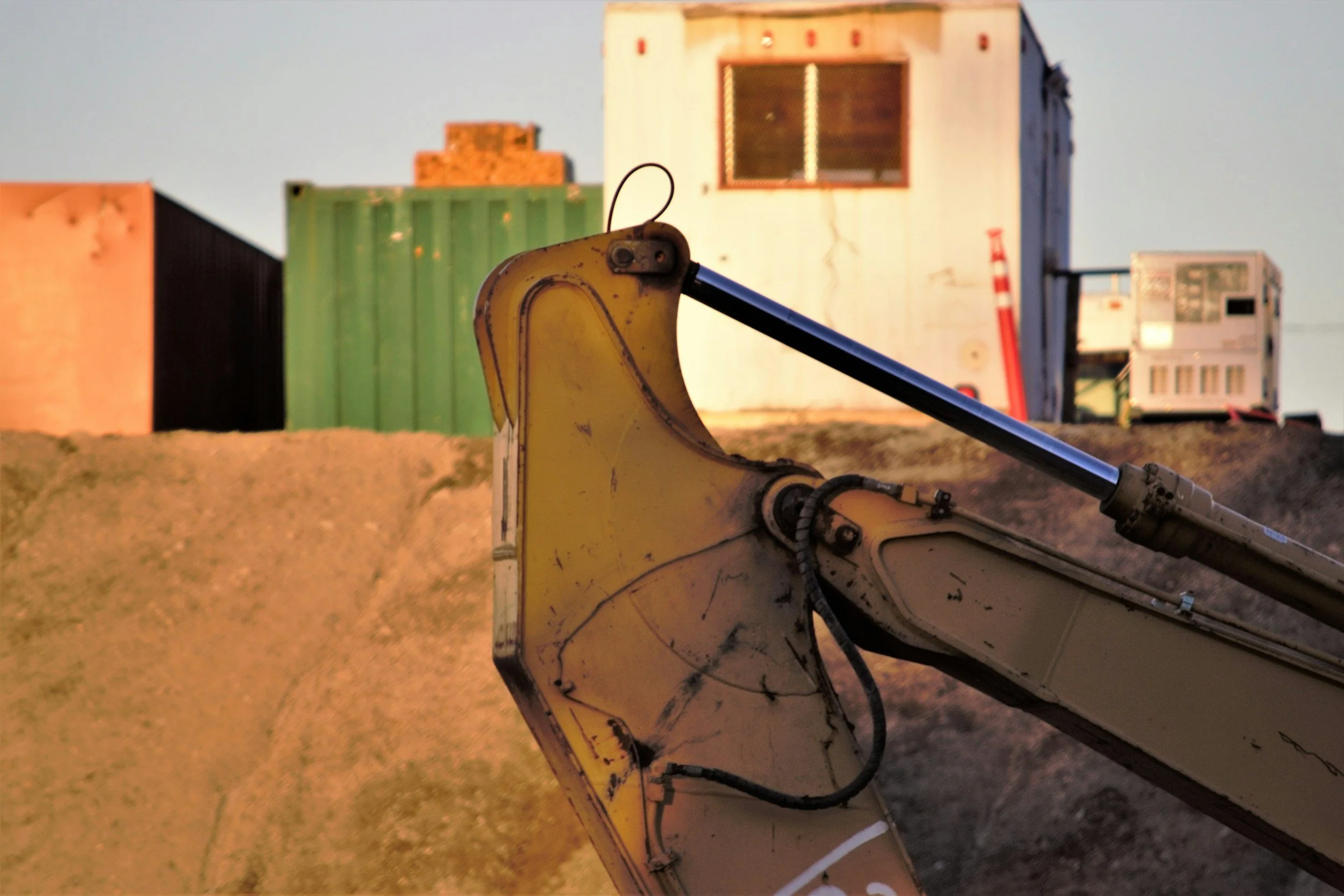 Close-up of a yellow construction or excavator arm with hydraulic hoses and metal parts, set against a background of colorful shipping containers and a building with windows.
