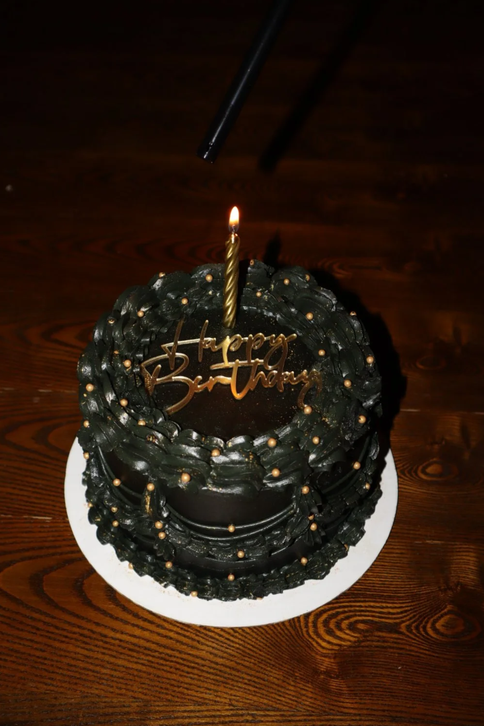 A black birthday cake decorated with gold beads and a gold "Happy Birthday" topper, with a lit single candle on top, placed on a white plate on a wooden table.