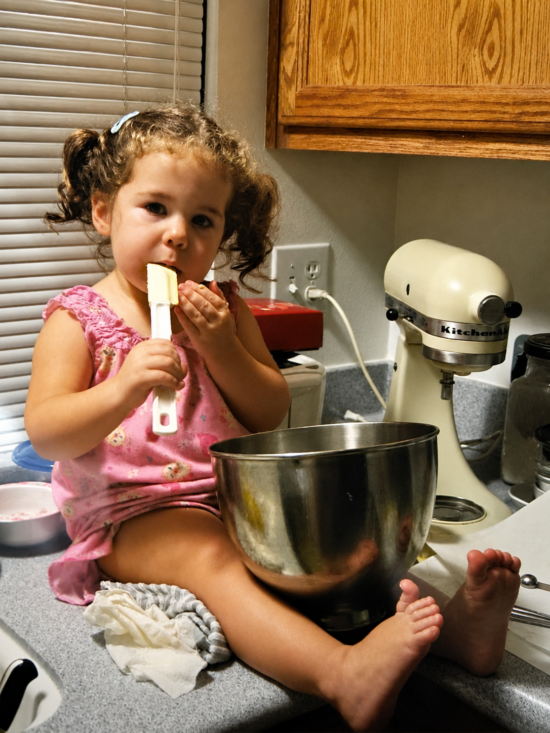 A young girl sitting on a kitchen counter, holding a spatula and licking it, with a large mixing bowl in front of her. She is wearing a pink dress with a pattern and has curly hair tied in pigtails.