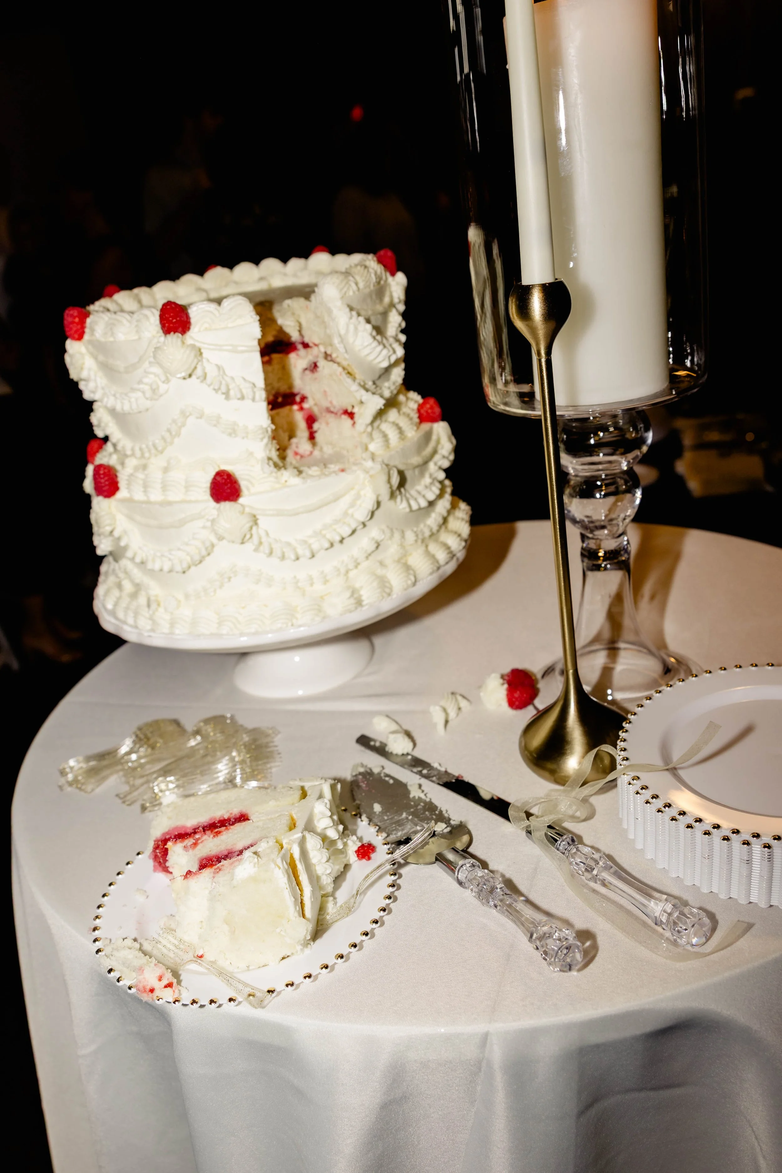 A white wedding cake with raspberry decorations and white icing, partially sliced on a white tablecloth. A slice of cake with raspberry filling and a plastic cake server is visible. A tall candle holder with tapered candles and a small round white cake with silver beads are on the table.