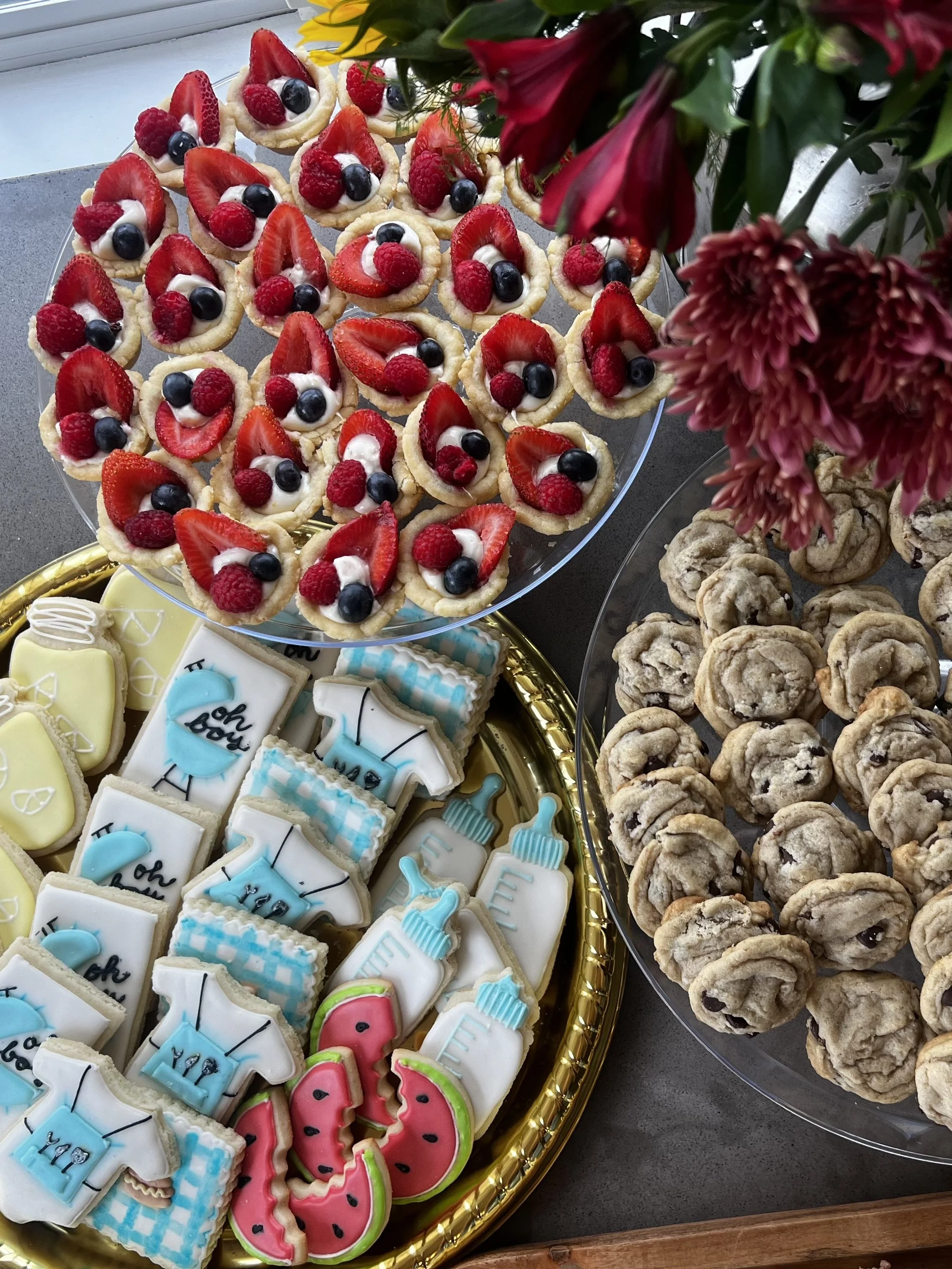 Tray with decorated cookies, including watermelon and birthday-themed cookies, and a plate of chocolate chip cookies with a bouquet of red flowers nearby.