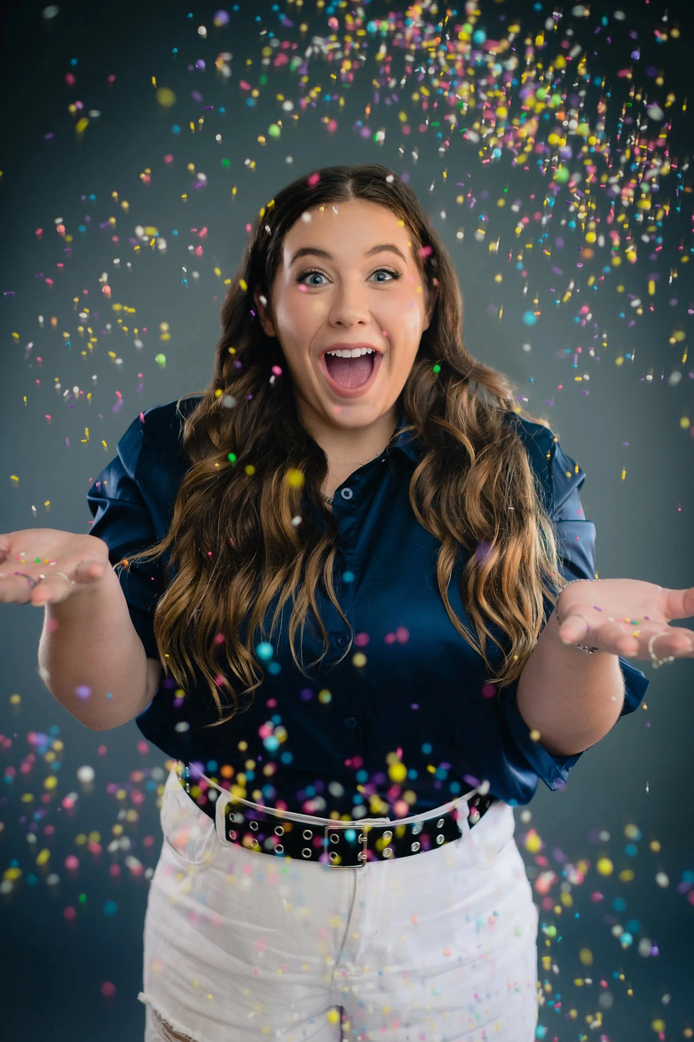 A woman with long brown hair and blue eyes celebrating with colorful confetti falling around her, smiling with her mouth open, wearing a dark blue shirt and light-colored shorts, against a dark background.