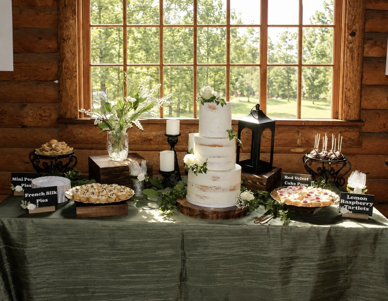 A dessert table with a three-tiered semi-naked white wedding cake, surrounded by floral arrangements, candles, and various dessert options including mini pecan pies, French silk pies, lemon raspberry tartlets, and red velvet cake pops, set against wooden walls and a large window overlooking a green outdoor scene.