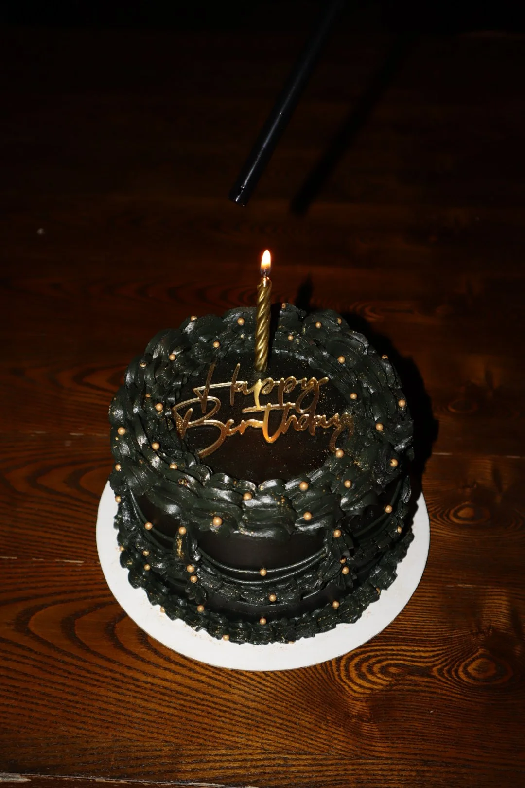 A black birthday cake with gold decorations and a gold candle, on a white plate, with a lit candle, on a wooden table.