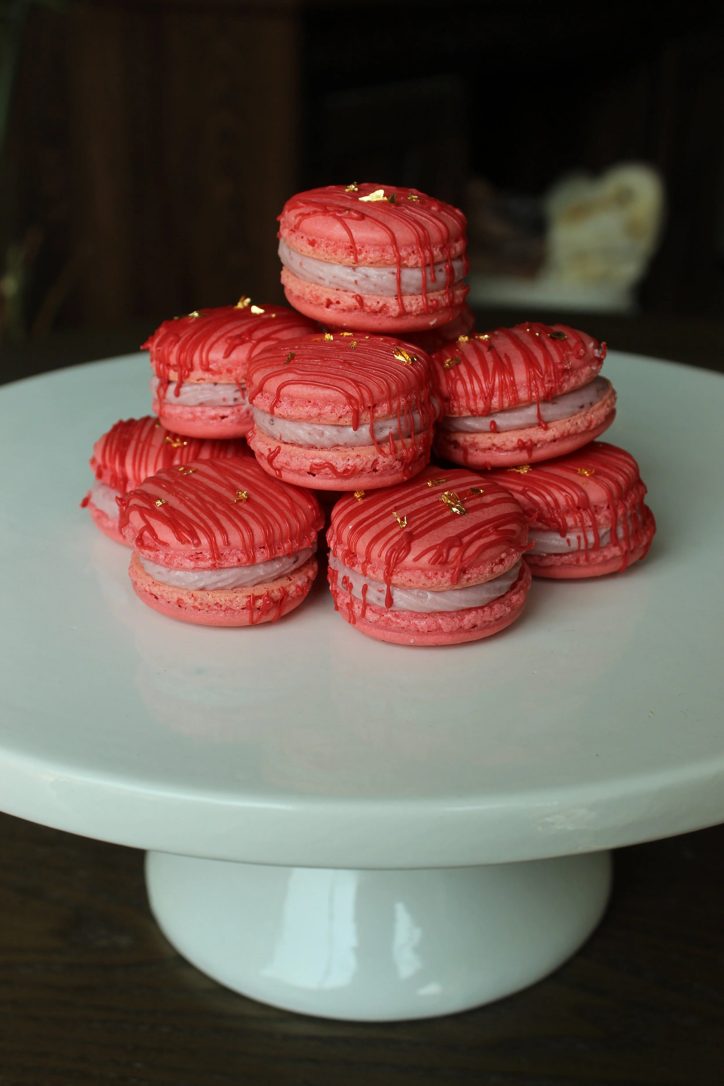 Pink macarons topped with red drizzle and gold leaf decorations on a white cake stand.