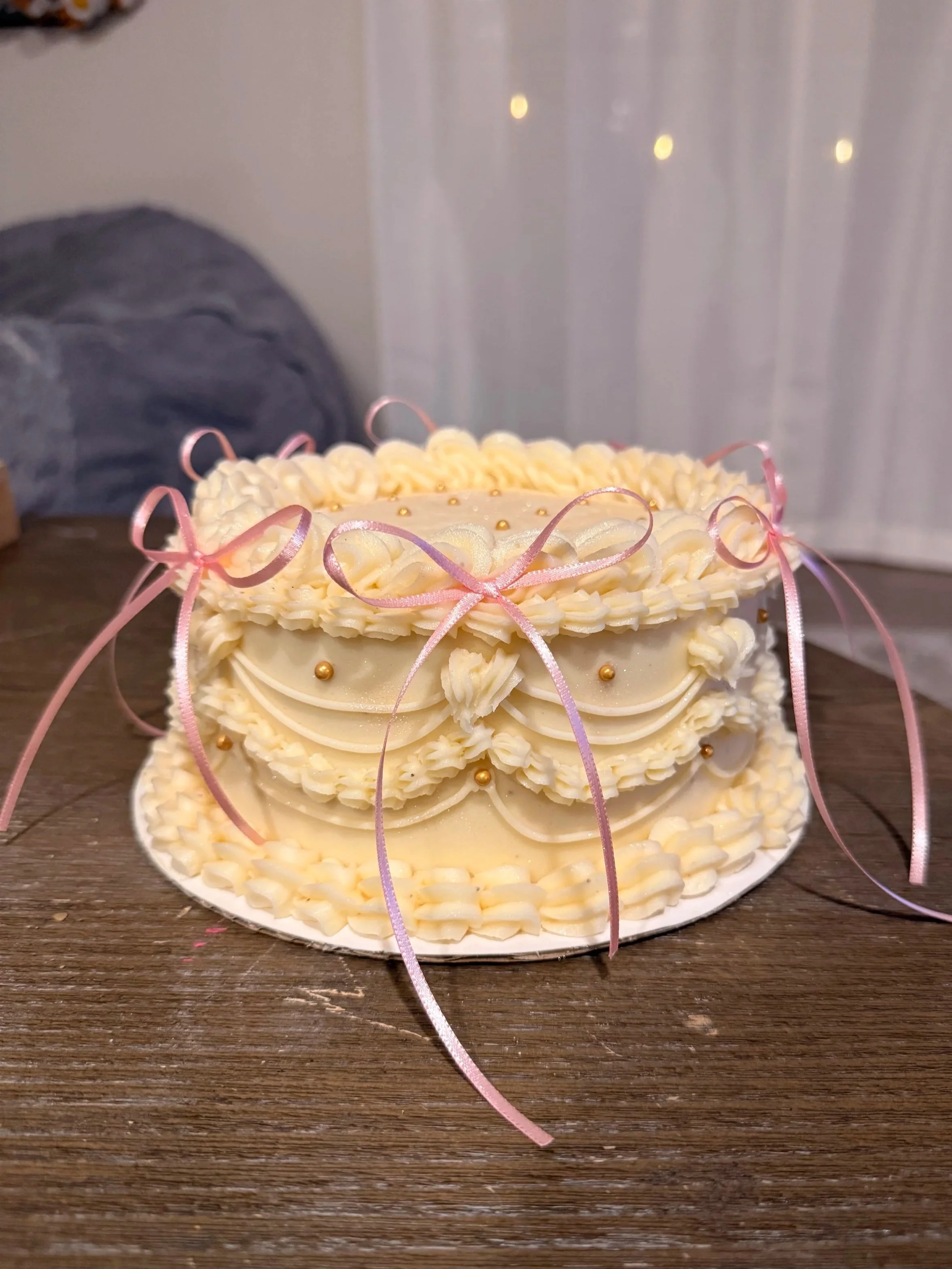 A layered white cake decorated with pink ribbons and gold beads, placed on a dark wooden surface with a blurred background.