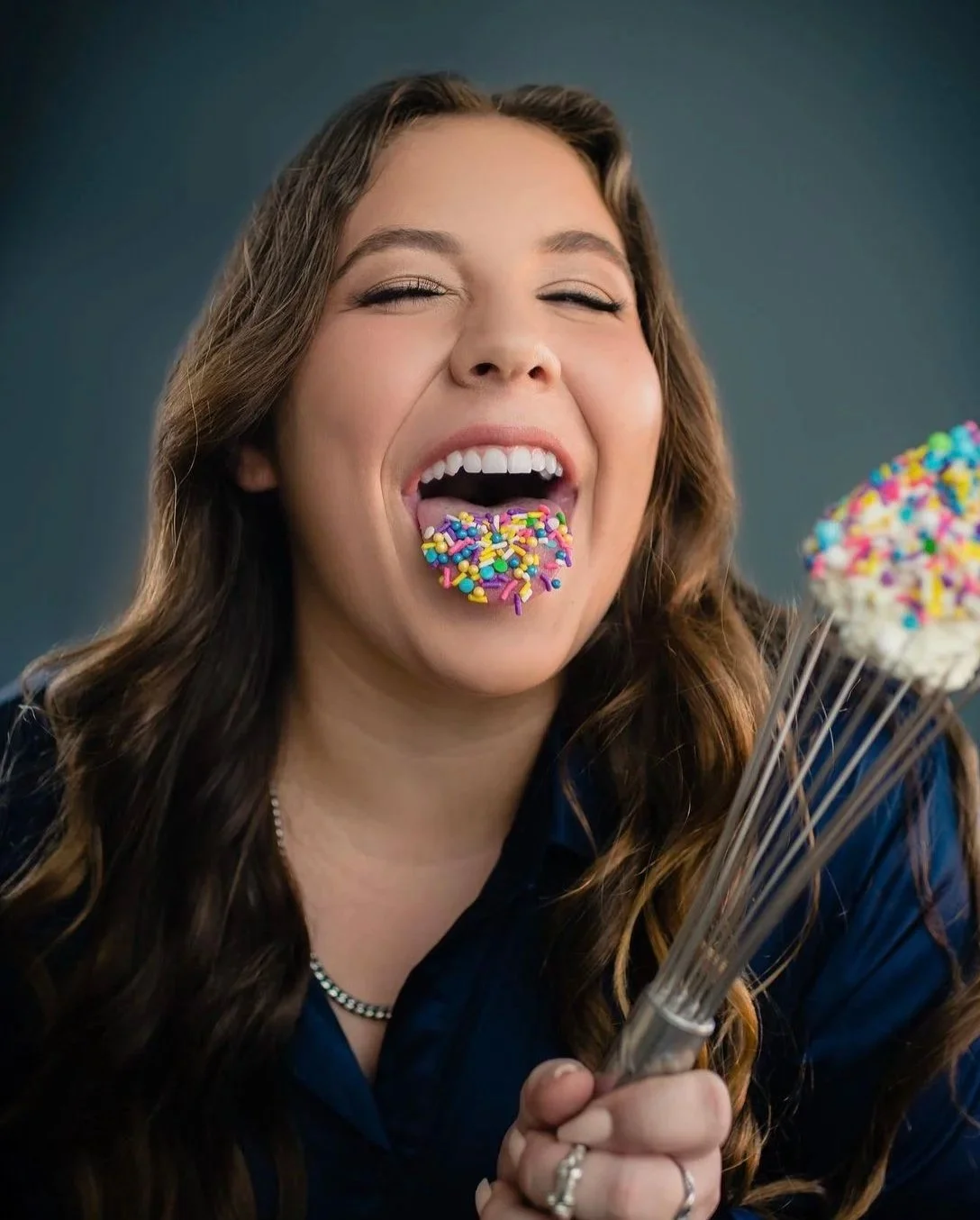 A woman with long wavy brown hair is smiling as she holds a wire whisk topped with white frosting and colorful sprinkles. She is sticking her tongue out with sprinkles on it, suggesting she is about to or has just enjoyed the sprinkles.