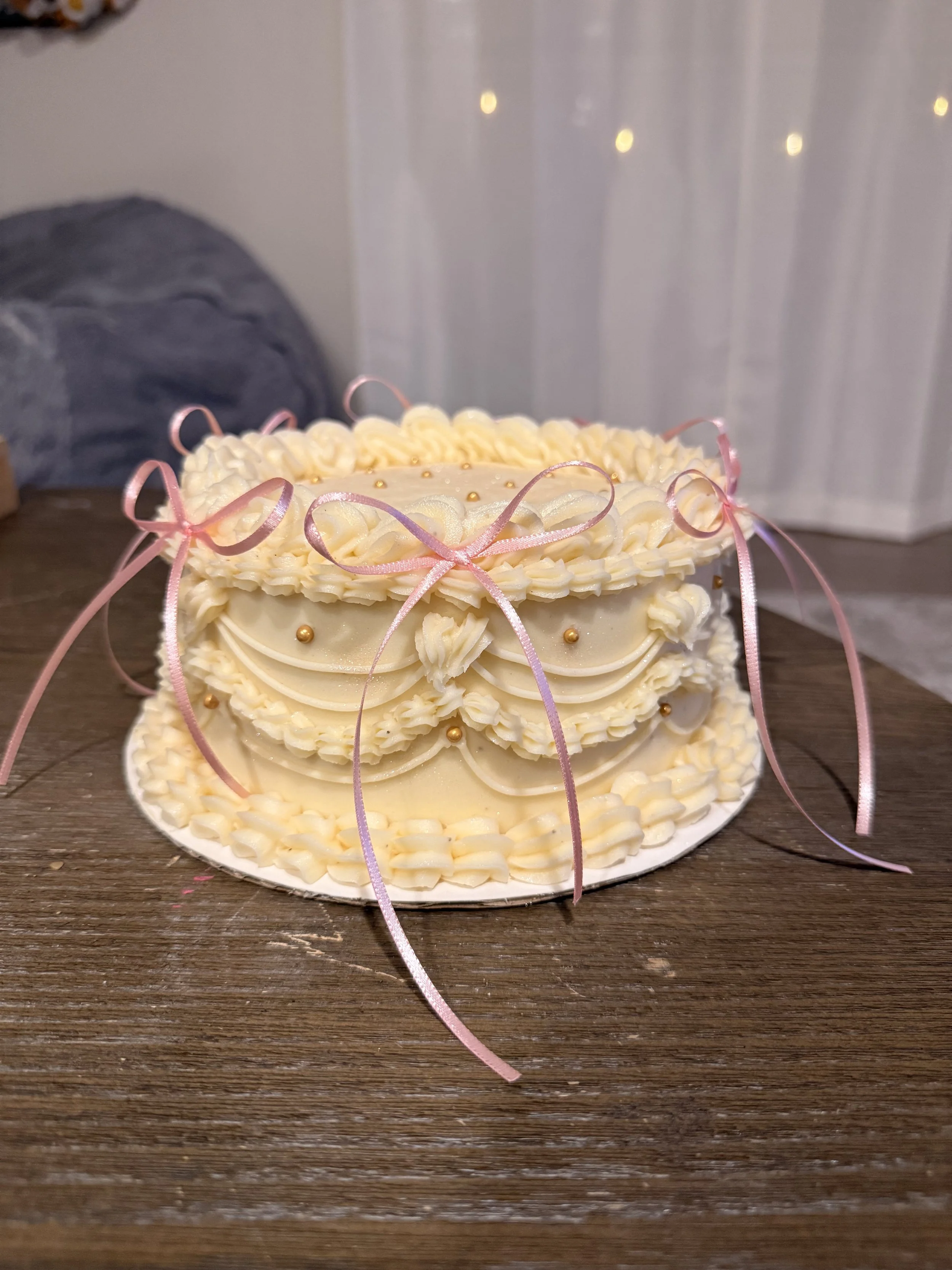 A round layered cake decorated with white cream, pink ribbons, and small gold beads, sitting on a wooden surface with a white background.