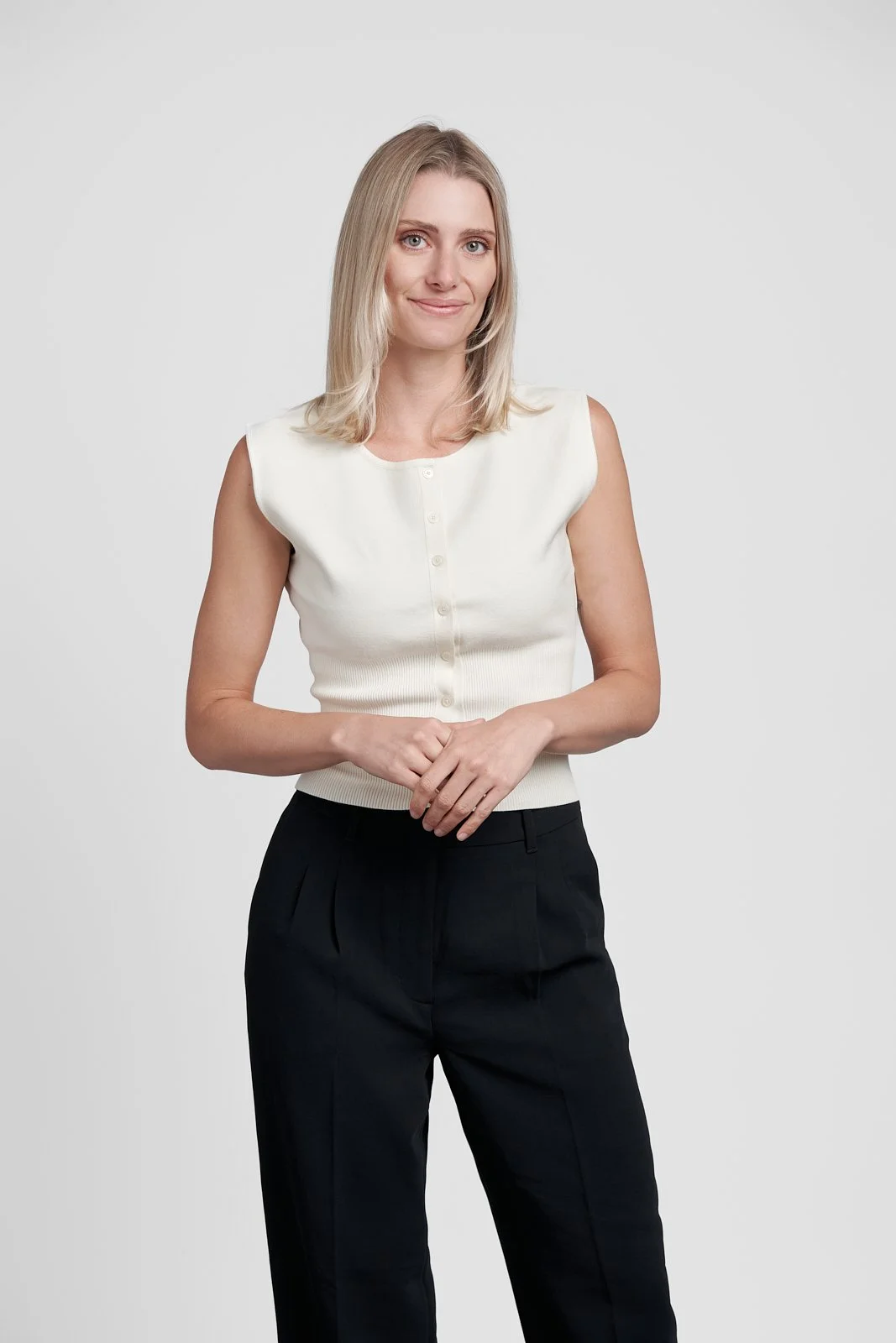 A young woman with blonde hair, wearing a sleeveless white button-up top and black pants, standing against a plain white background.