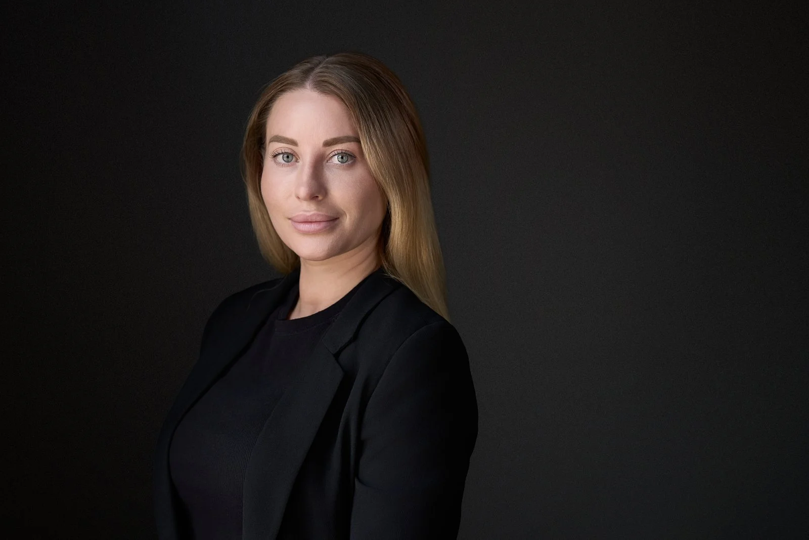 A woman with long blonde hair, fair skin, and blue eyes, wearing a black blazer and sitting against a dark background.