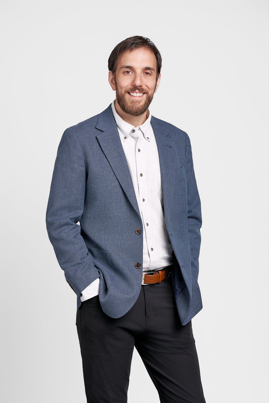A man with a beard and mustache, dressed in a blue blazer, white shirt, and dark pants, standing with one hand in his pocket and smiling against a plain white background.