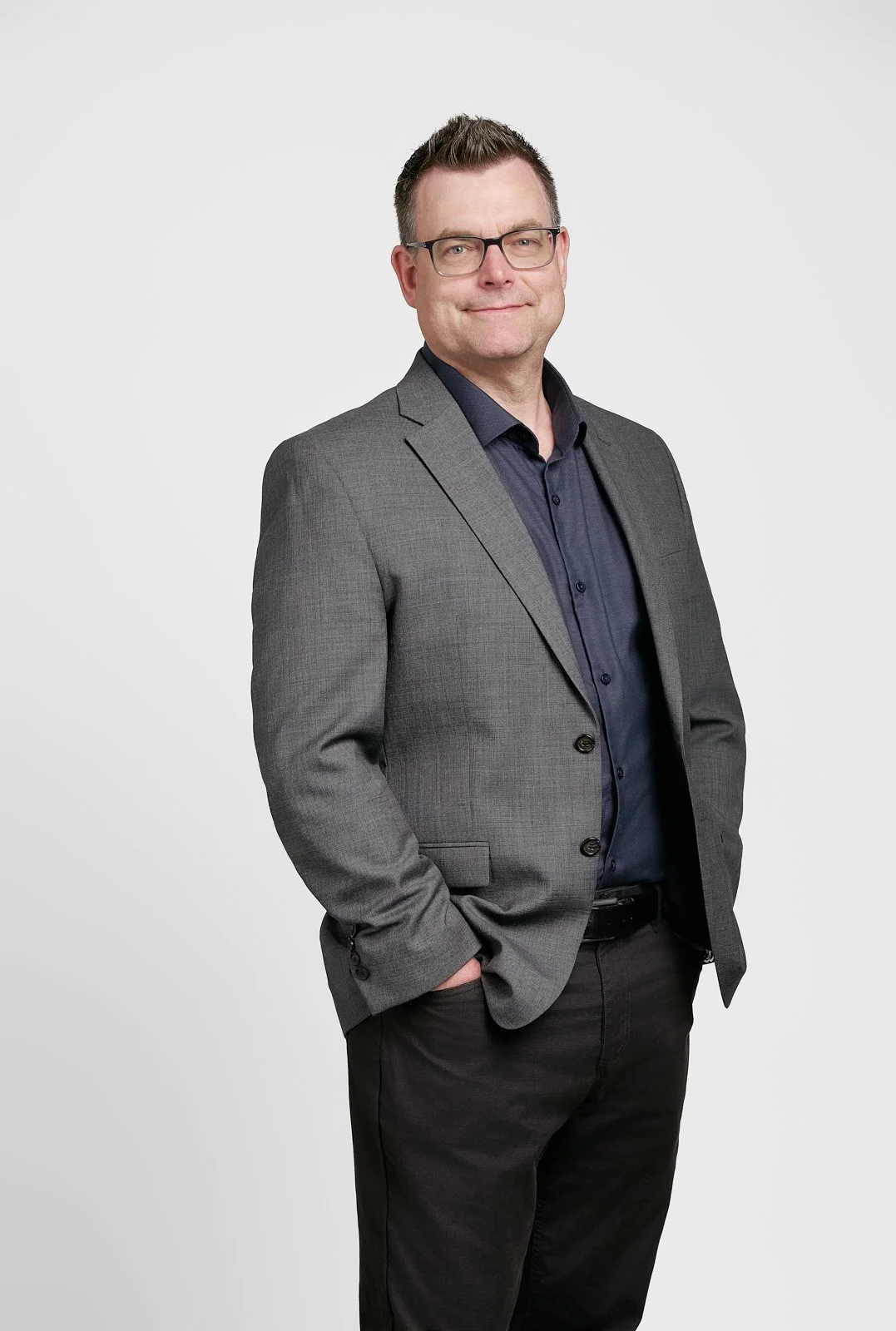 A man in a gray suit and black shirt standing against a white background with his hand in his pocket, smiling slightly at the camera.