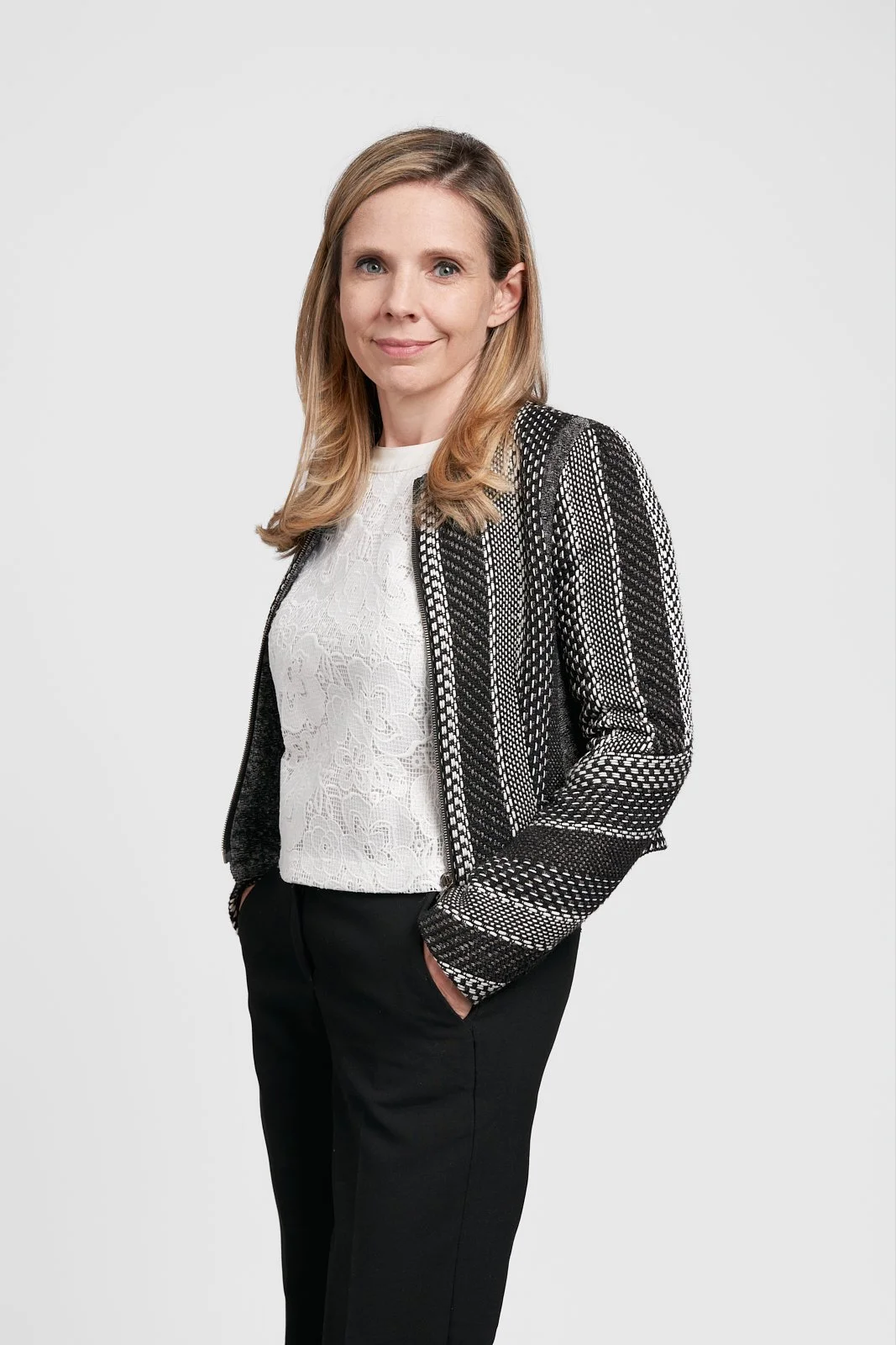 A confident woman with shoulder-length blonde hair, wearing a white lace blouse, black pants, and a black and white patterned jacket, standing with one hand in her pocket against a plain light gray background.