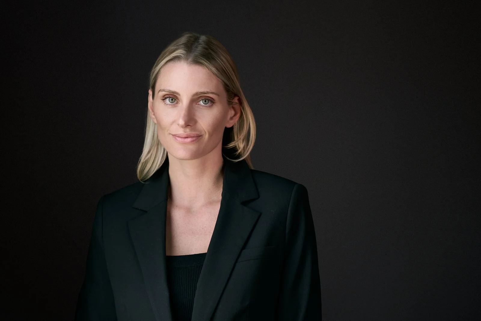 A woman with blonde hair in a black blazer against a dark background.
