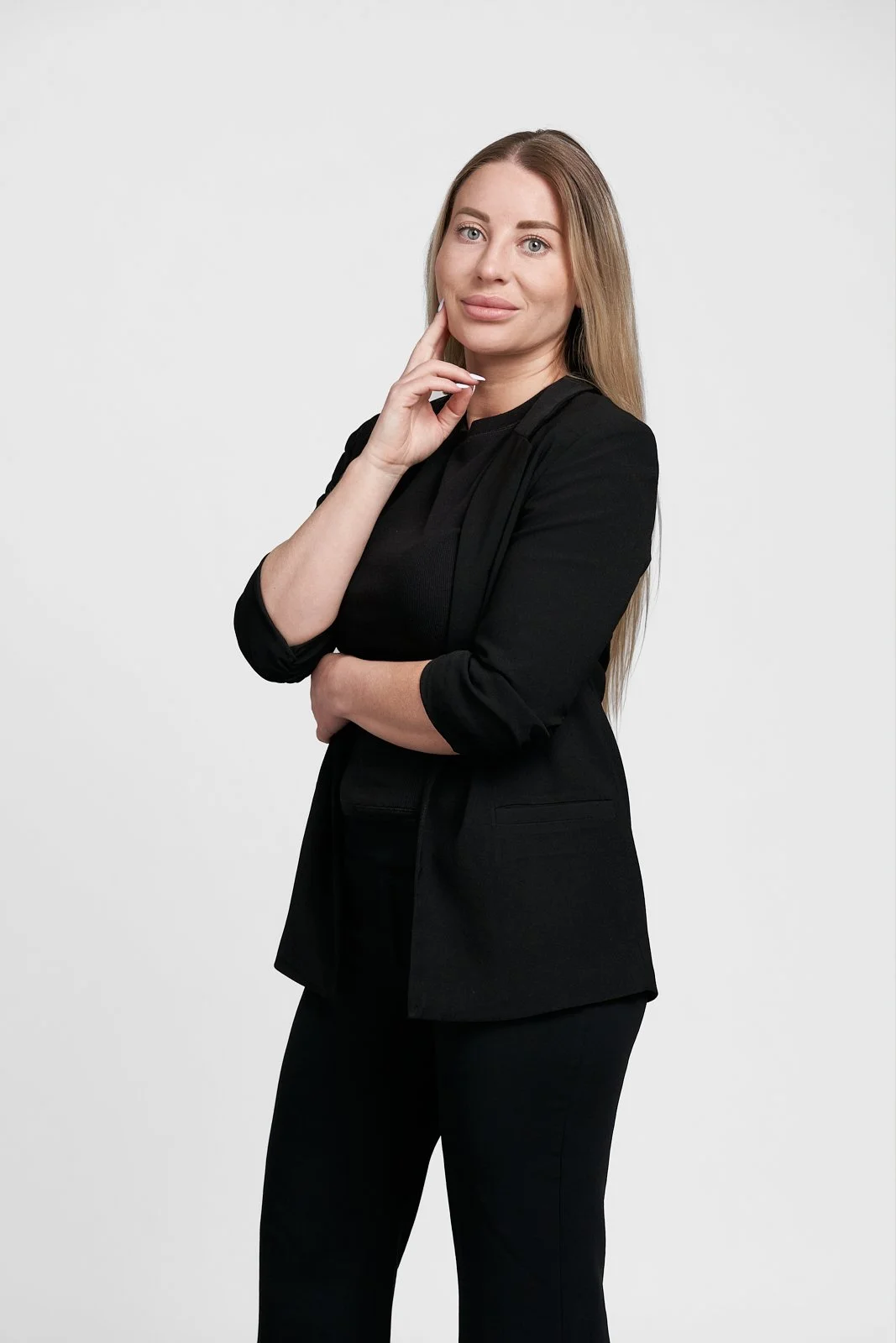 A woman with long blonde hair wearing a black blazer and black pants, standing with her left arm crossed and her right hand touching her face against a plain white background.