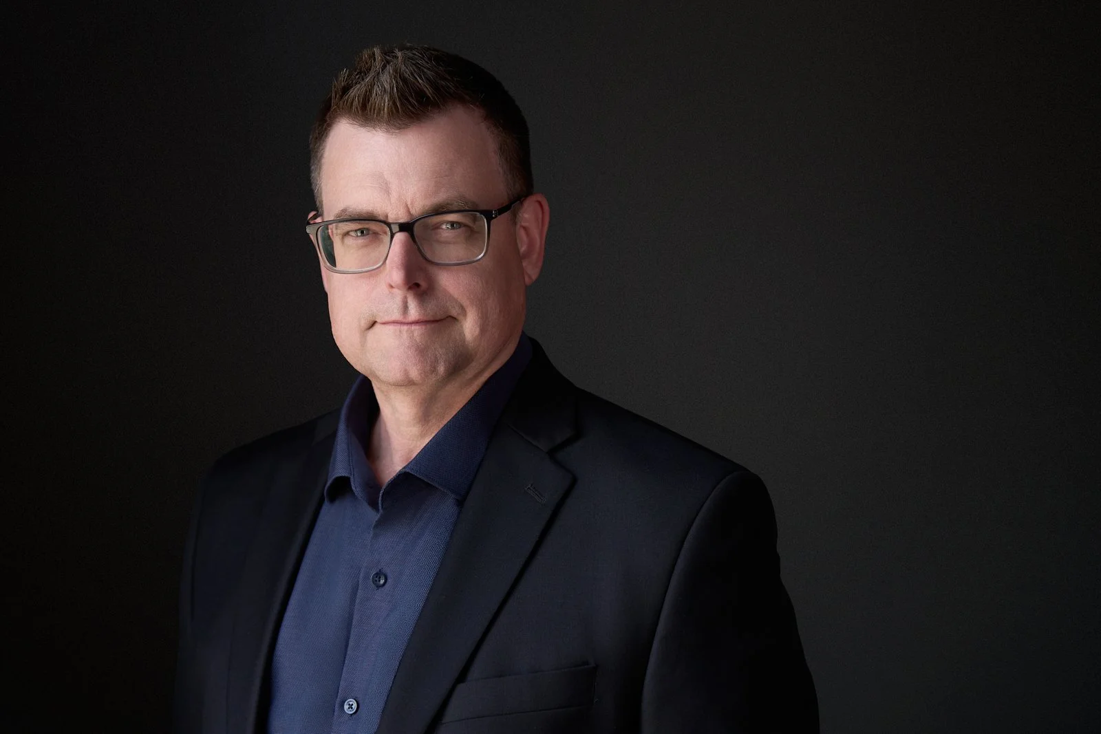 A middle-aged man with short brown hair and glasses, wearing a dark suit jacket over a blue collared shirt, posing against a dark background.