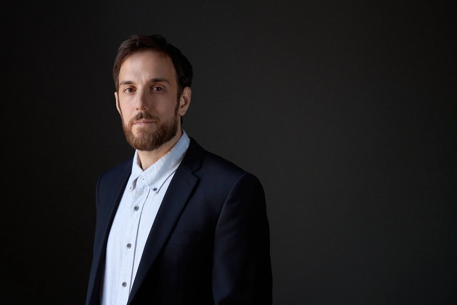 Portrait of a young man with a beard and short brown hair, wearing a dark suit jacket and a light-colored button-up shirt, standing against a dark background.