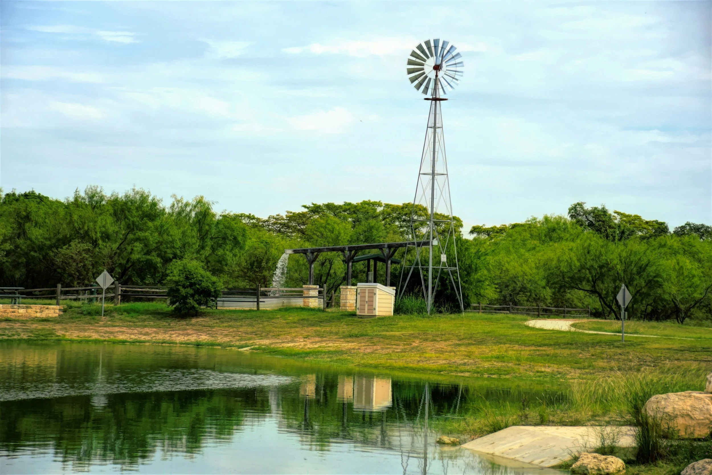 A windmill beside a lake with lush green trees in the background and a partly cloudy sky.