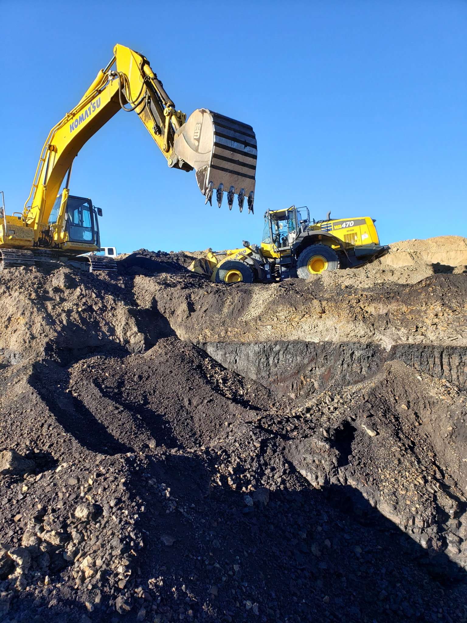 A yellow excavator with a metal bucket is digging into dirt and rocks on a construction site under a clear blue sky.