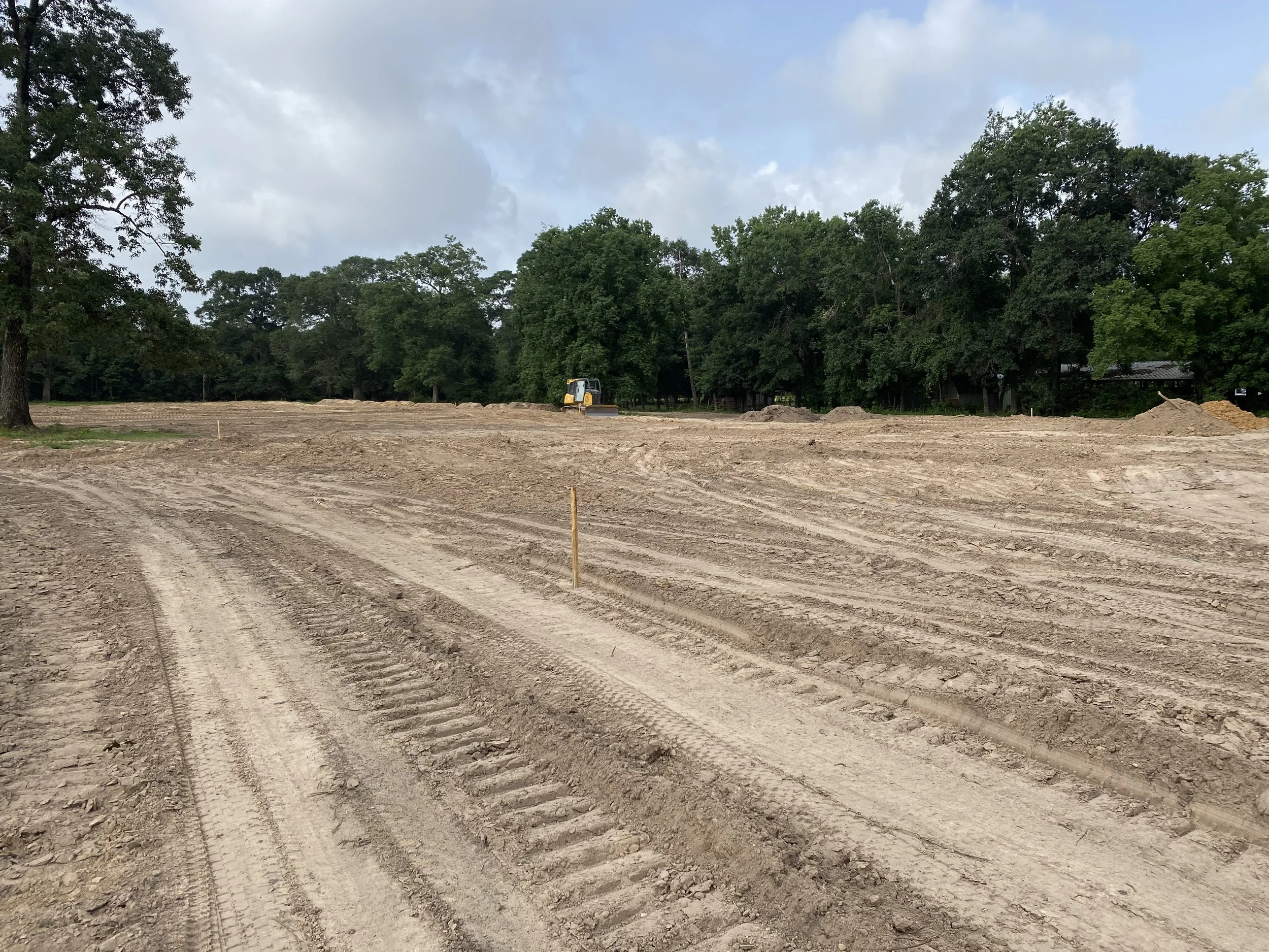 Construction site with a dirt ground, tire tracks, and a small excavator surrounded by trees and cloudy sky.