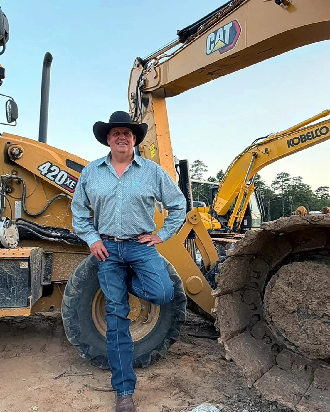 A man wearing a black cowboy hat, a light blue checkered shirt, and blue jeans stands in front of yellow construction equipment, including a Caterpillar bulldozer and a Kobelco excavator, at a construction site with dirt ground and trees in the background.