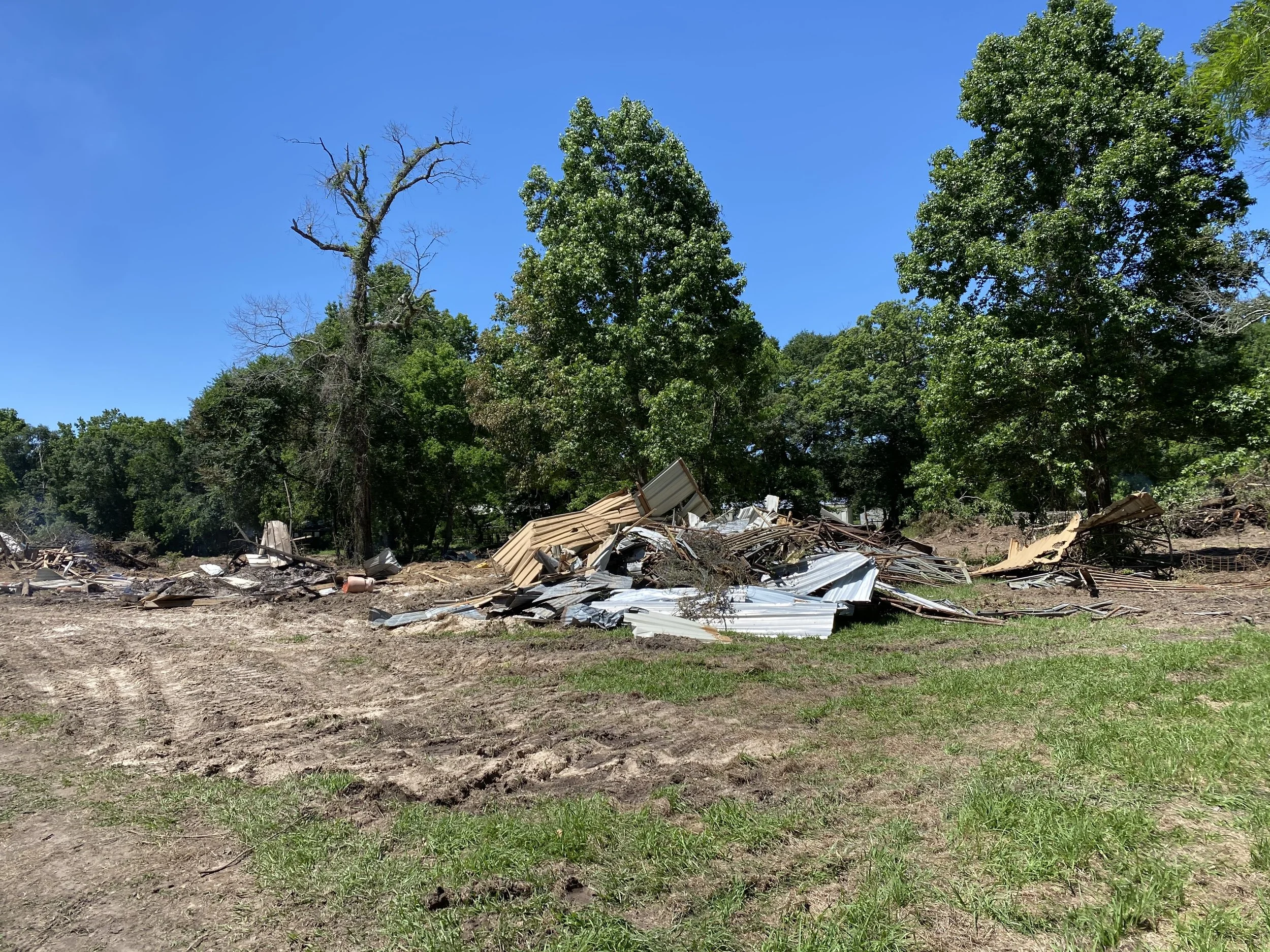 Debris and collapsed metal sheets on the ground with green trees and a bright blue sky in the background.