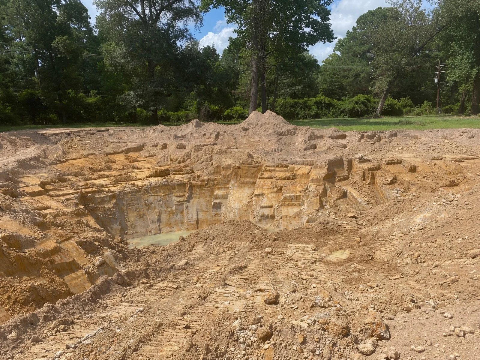 A large excavation site with exposed layered earth, dirt, and rocks, surrounded by green trees and a partly cloudy sky.