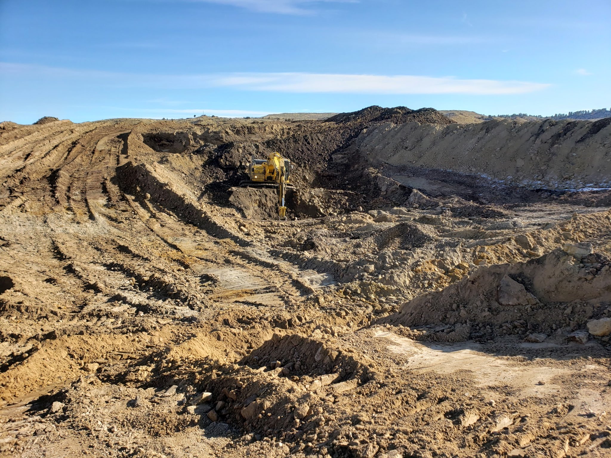 A construction site with a yellow excavator digging into a large mound of dirt, with tire tracks visible in the foreground and a clear blue sky above.