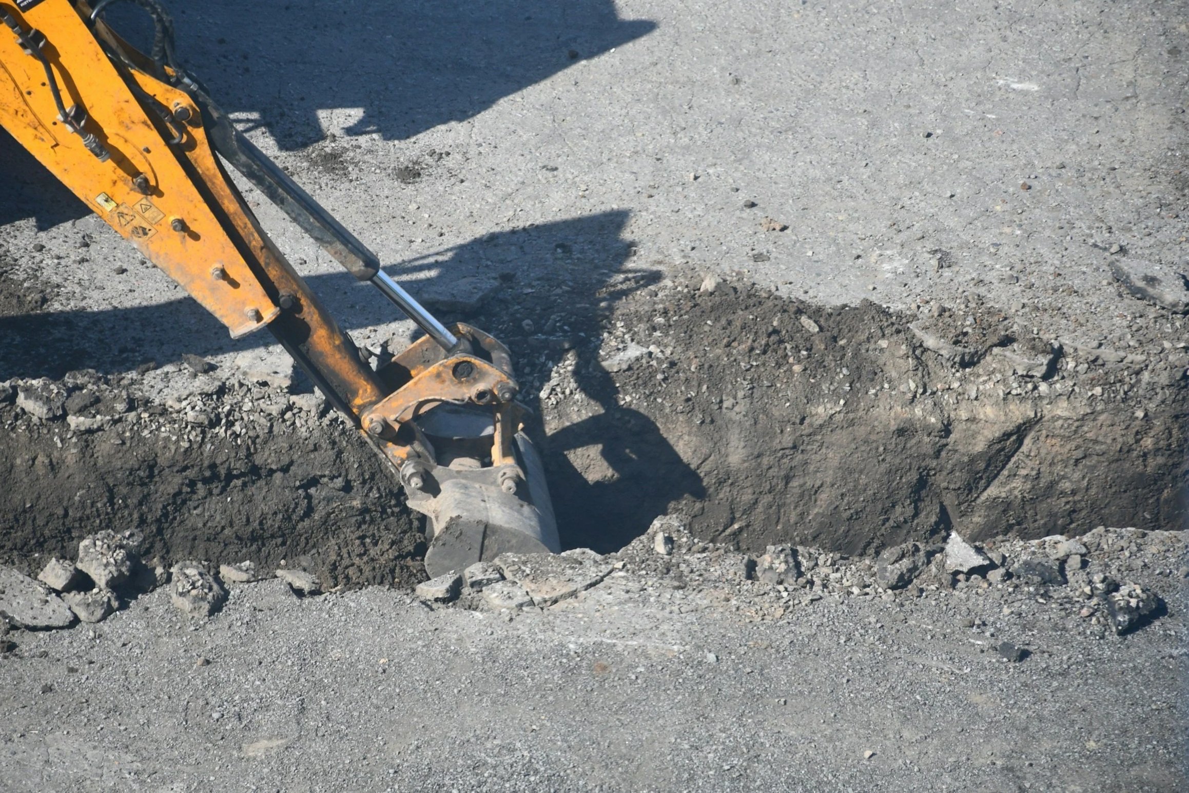 Close-up of a yellow excavator arm working on a dirt road, with disturbed soil and loose gravel.