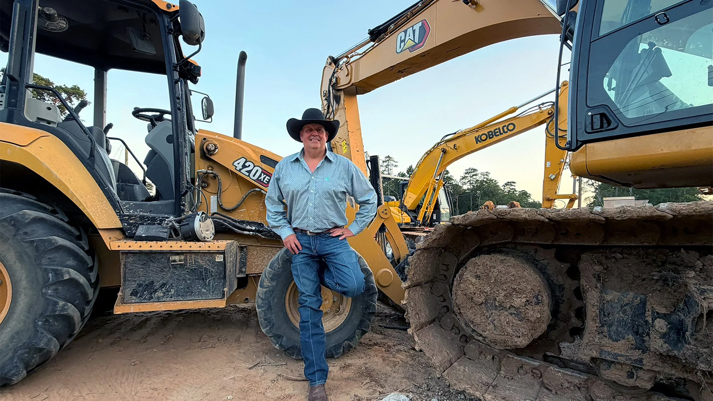 A man wearing a cowboy hat and blue jeans standing on a dirt ground next to large yellow construction vehicles, including an excavator and a loader, in an outdoor construction site during daylight.