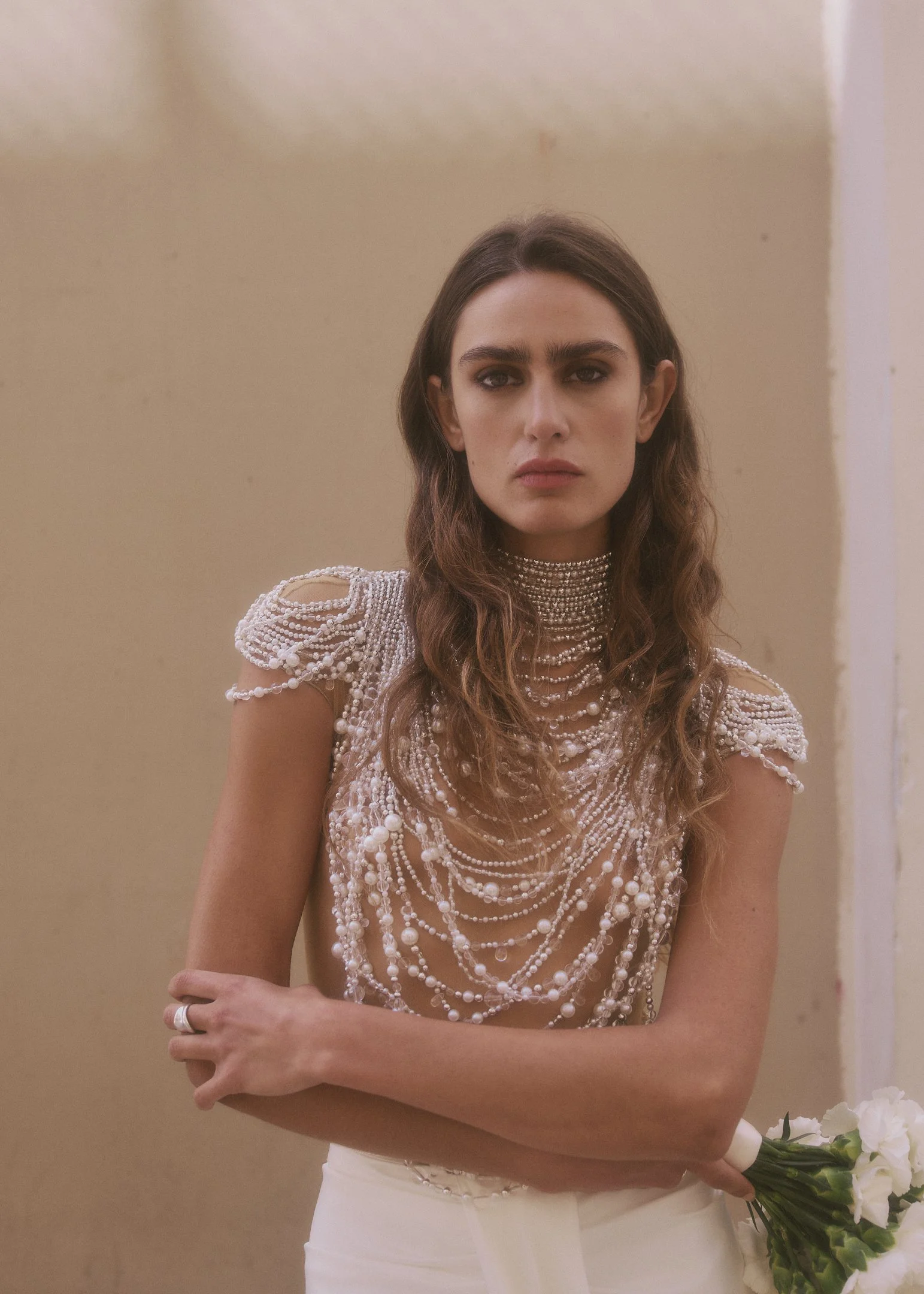 Elena with arms crossed and a white bouquet against a sunlit beige wall, bridal portrait near Barcelona.