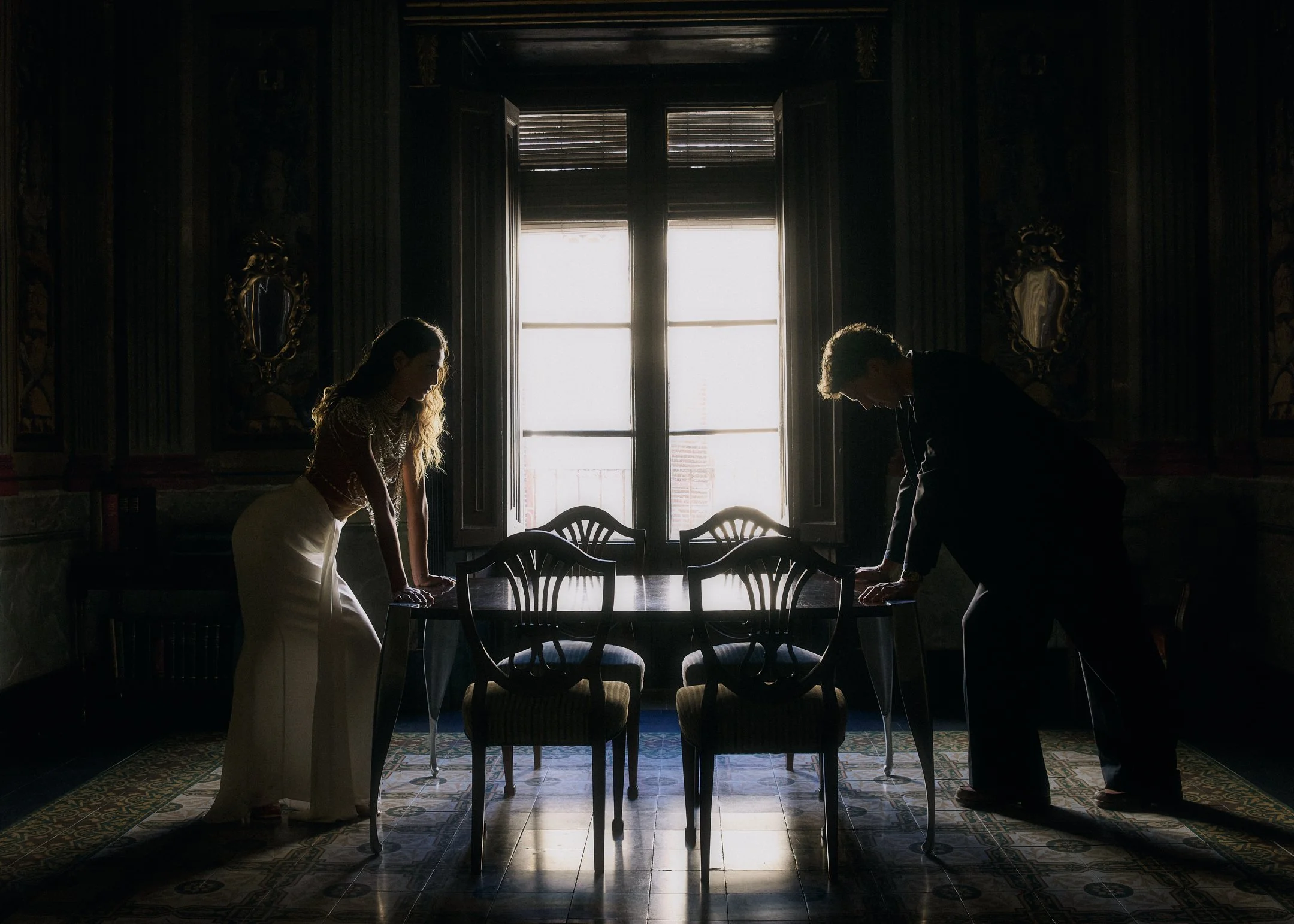 Elena and Oscar in silhouette, leaning over a dining table before a tall shuttered window in a historic salon.