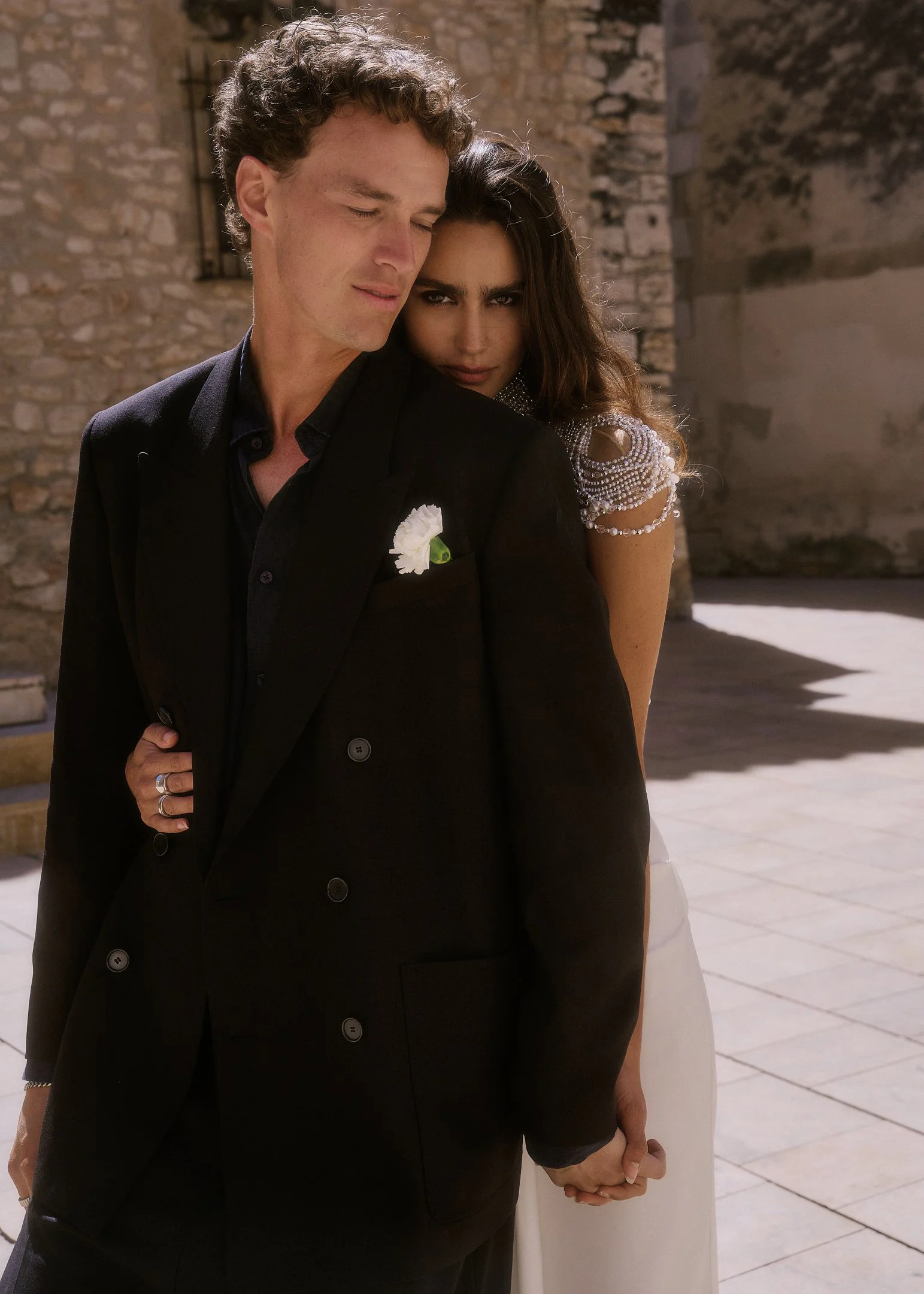 Elena embracing Oscar from behind in strong Mediterranean sunlight, destination wedding near Barcelona.