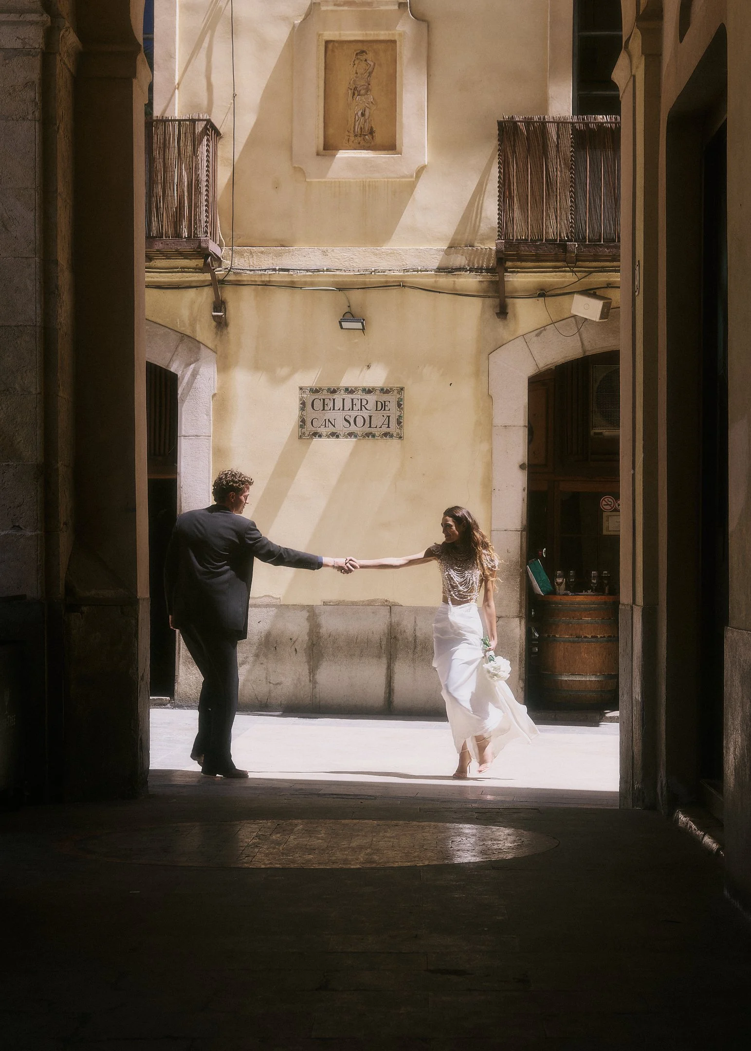 Elena and Oscar holding hands in a sunlit street, seen through a shaded passage in an old Catalan town.