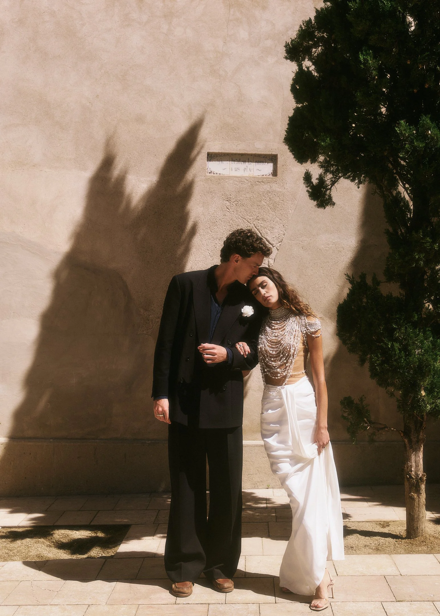 Elena resting her head on Oscar’s shoulder beside a cypress tree and its shadow on a warm wall in Spain.
