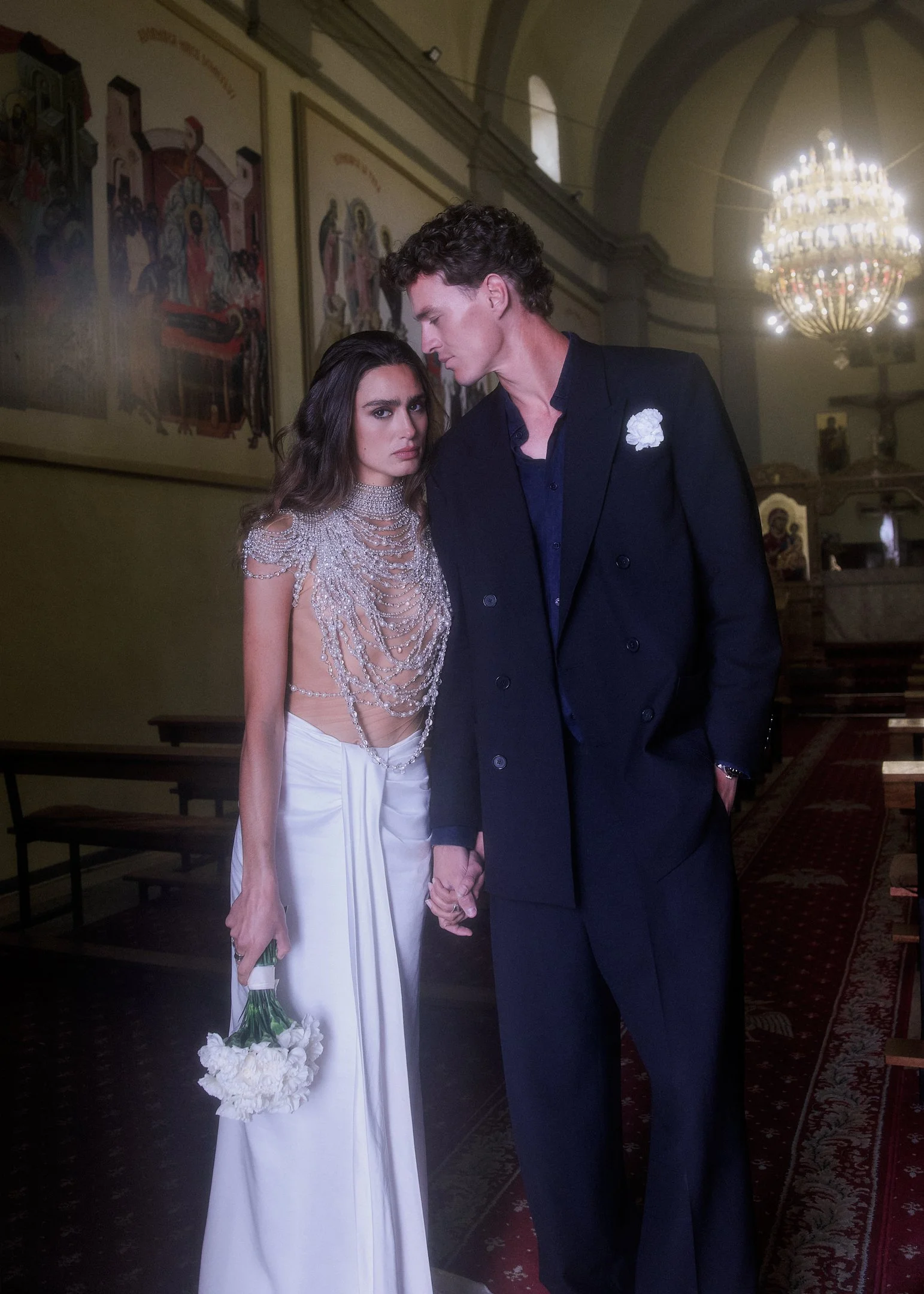 Elena and Oscar holding hands in a church aisle beneath a chandelier, wedding ceremony near Barcelona.