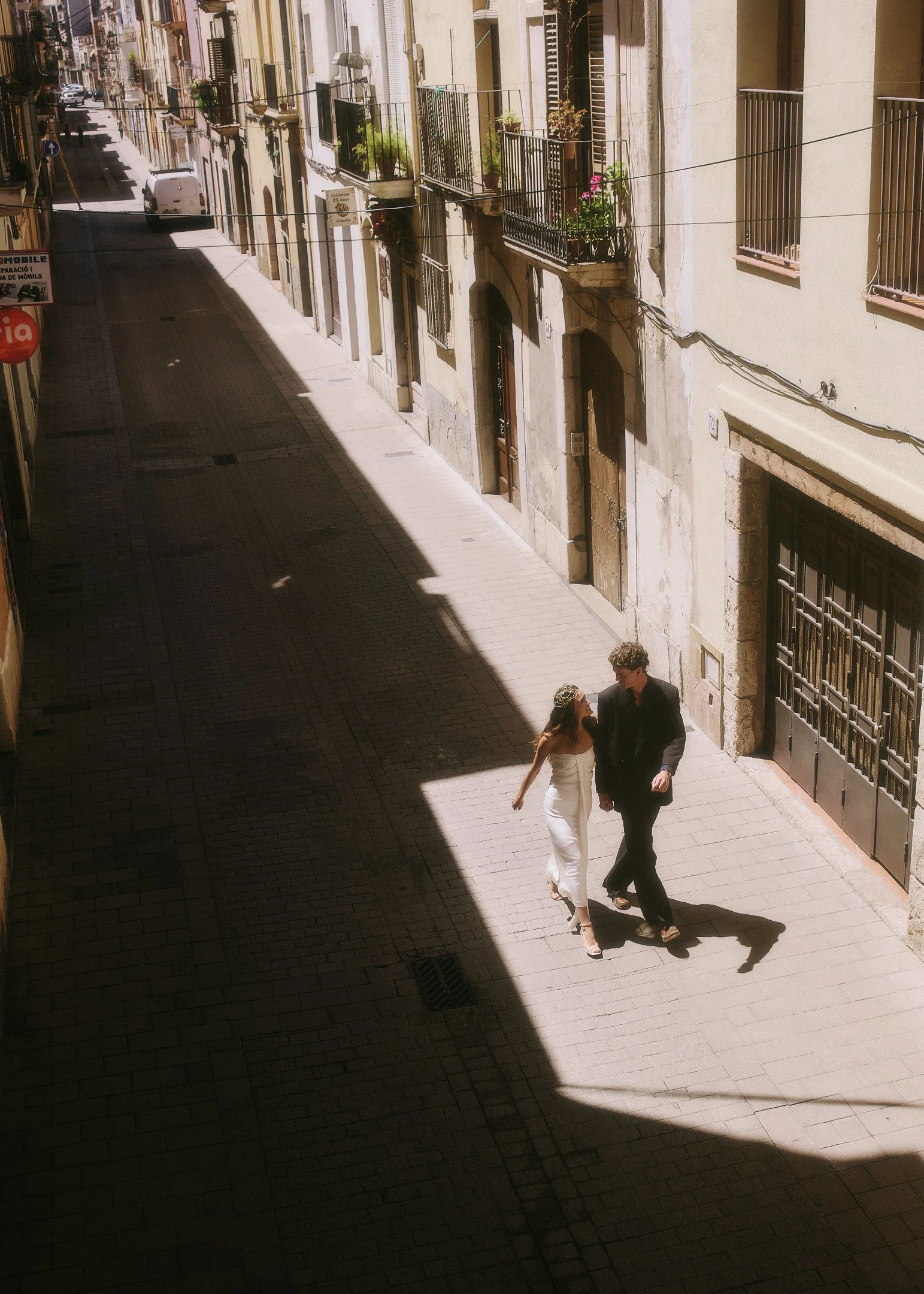 Elena and Oscar walking hand in hand down a sunlit street near Barcelona, photographed from above.