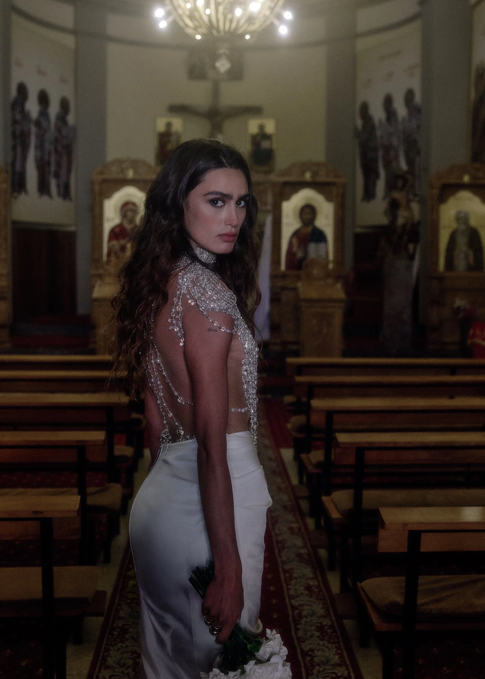 Elena looking over her shoulder in a church with icons and wooden pews, holding white carnations.