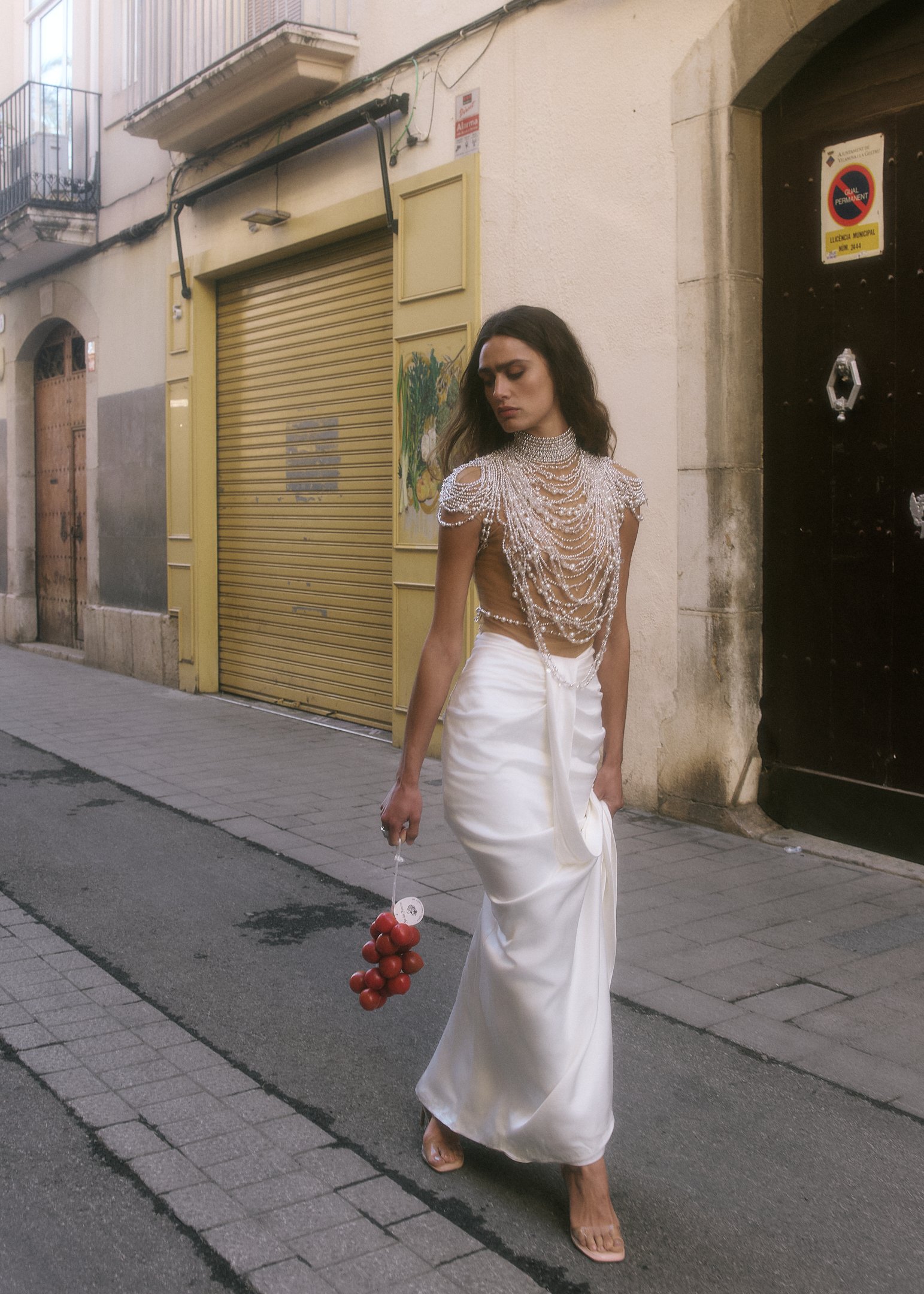 Elena in a pearl-embellished top and white satin skirt walking through an old town street near Barcelona, carrying red tomatoes.