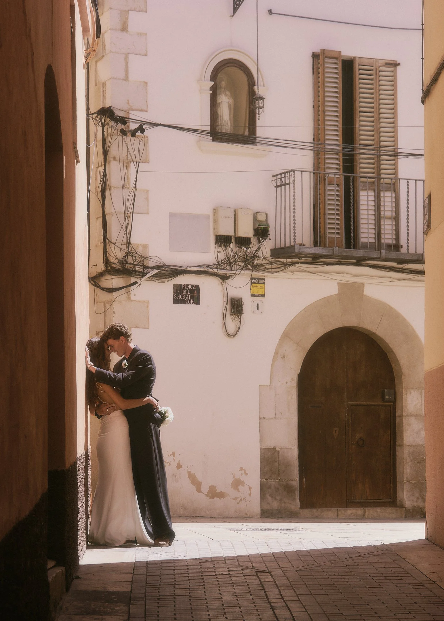 Elena and Oscar embracing in a narrow sunlit street in front of an arched wooden door in Catalonia.