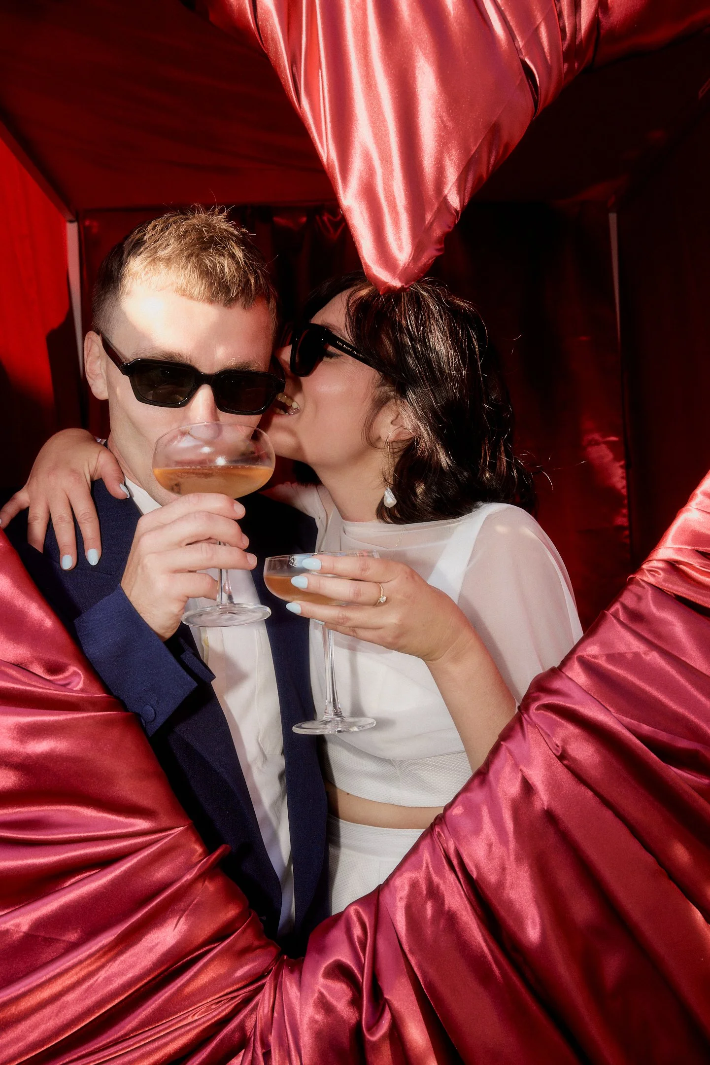 Braut küsst den Bräutigam mit Sonnenbrille vor roten Stoffbahnen im Hallo Emil Stuttgart, beide halten Cocktailschalen.