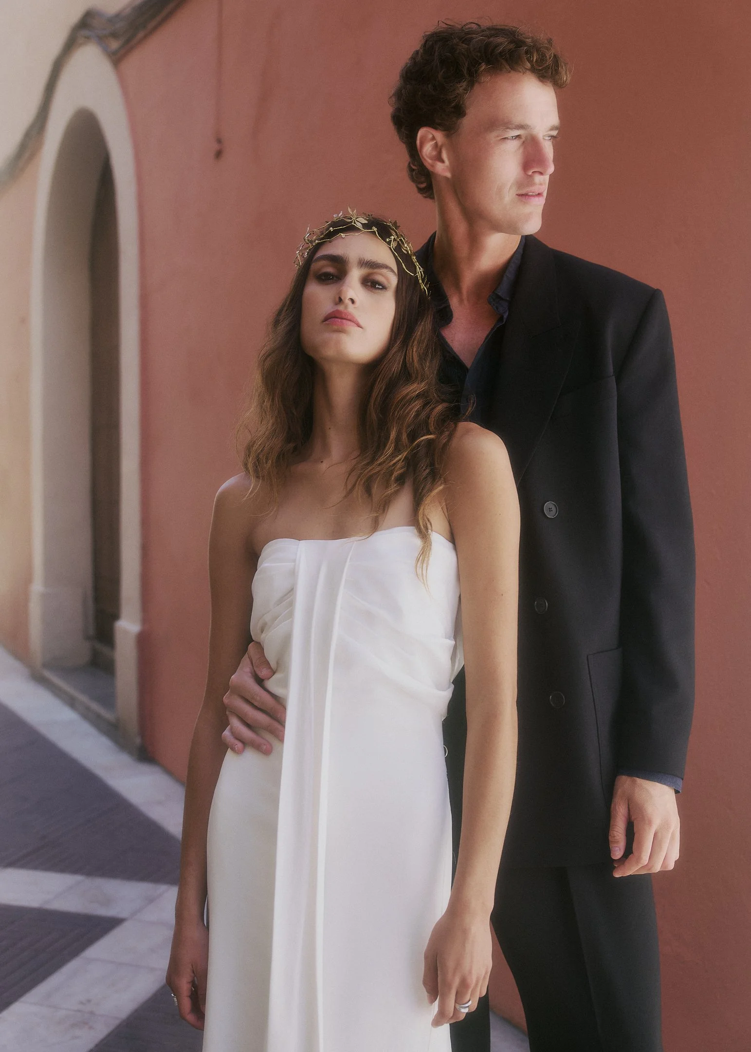 Elena in a strapless white Clio Peppiatt gown and golden headpiece, standing with Oscar in front of a terracotta wall.