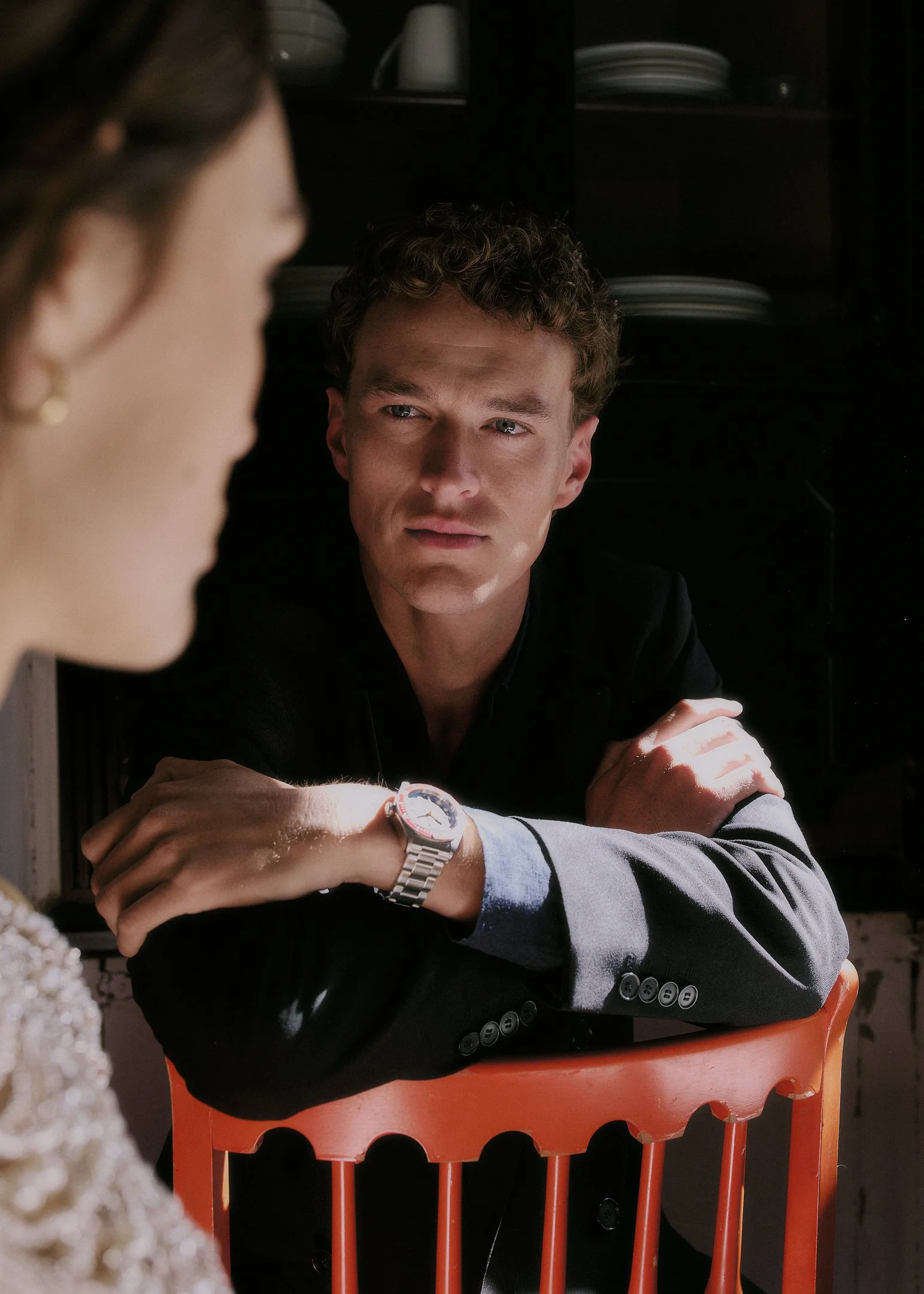 Oscar leaning on a red wooden chair and looking at Elena in a townhouse kitchen near Barcelona.
