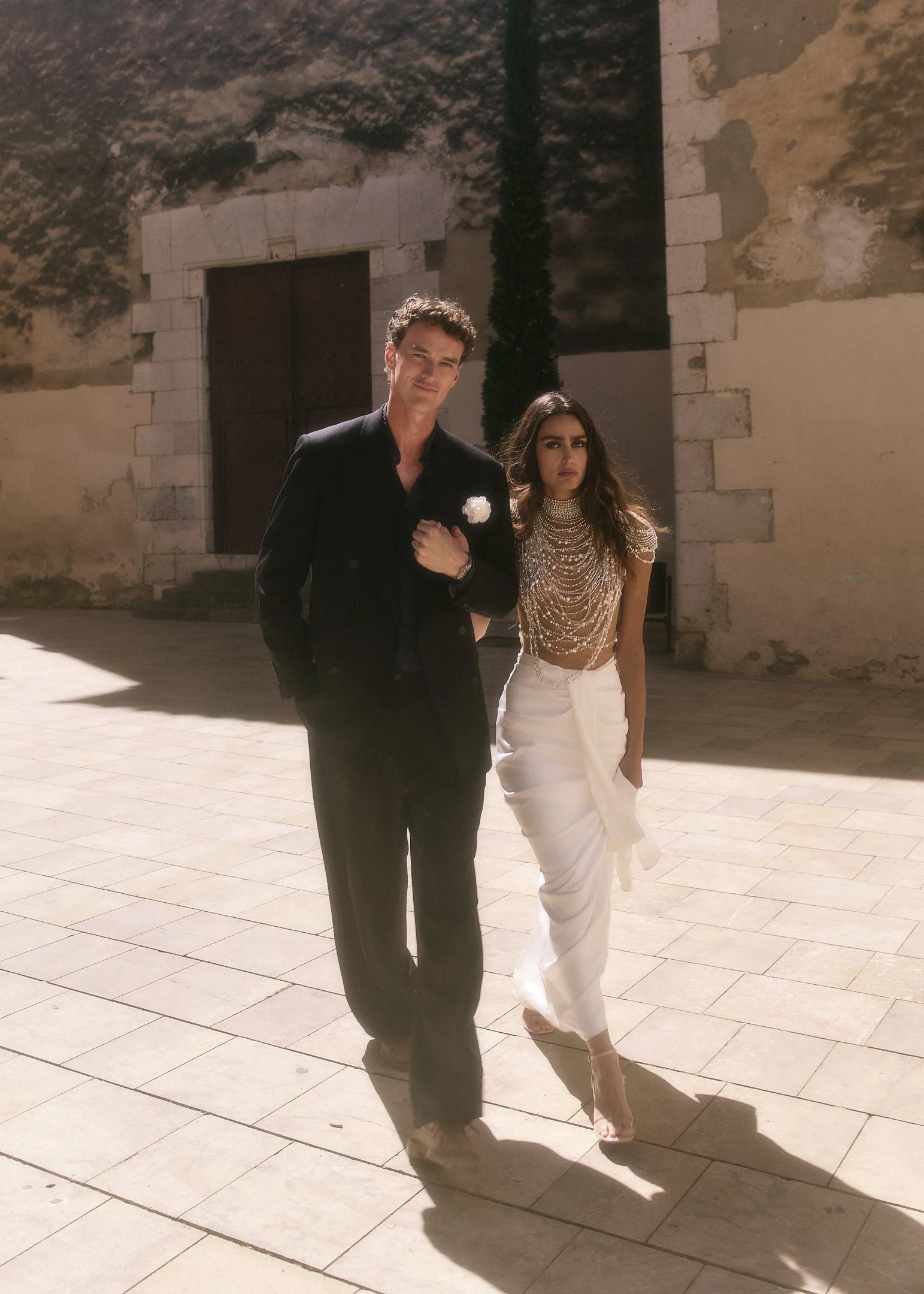 Elena and Oscar walking arm in arm across a sunlit square, wedding portrait in Spain.