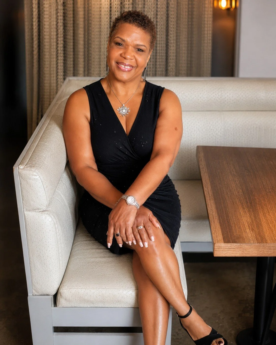 A woman sitting on a cream-colored booth in a restaurant, wearing a black dress, silver jewelry, and black shoes, smiling at the camera.