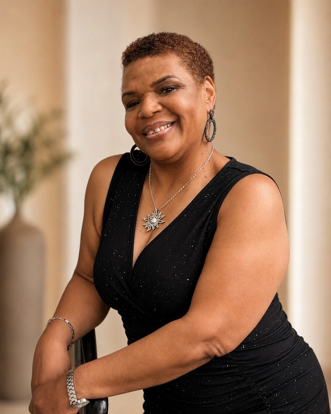 A woman with short curly hair smiling, wearing a black dress with sparkles, a necklace with a floral design, hoop earrings, and a bracelet, standing indoors with a blurred background.
