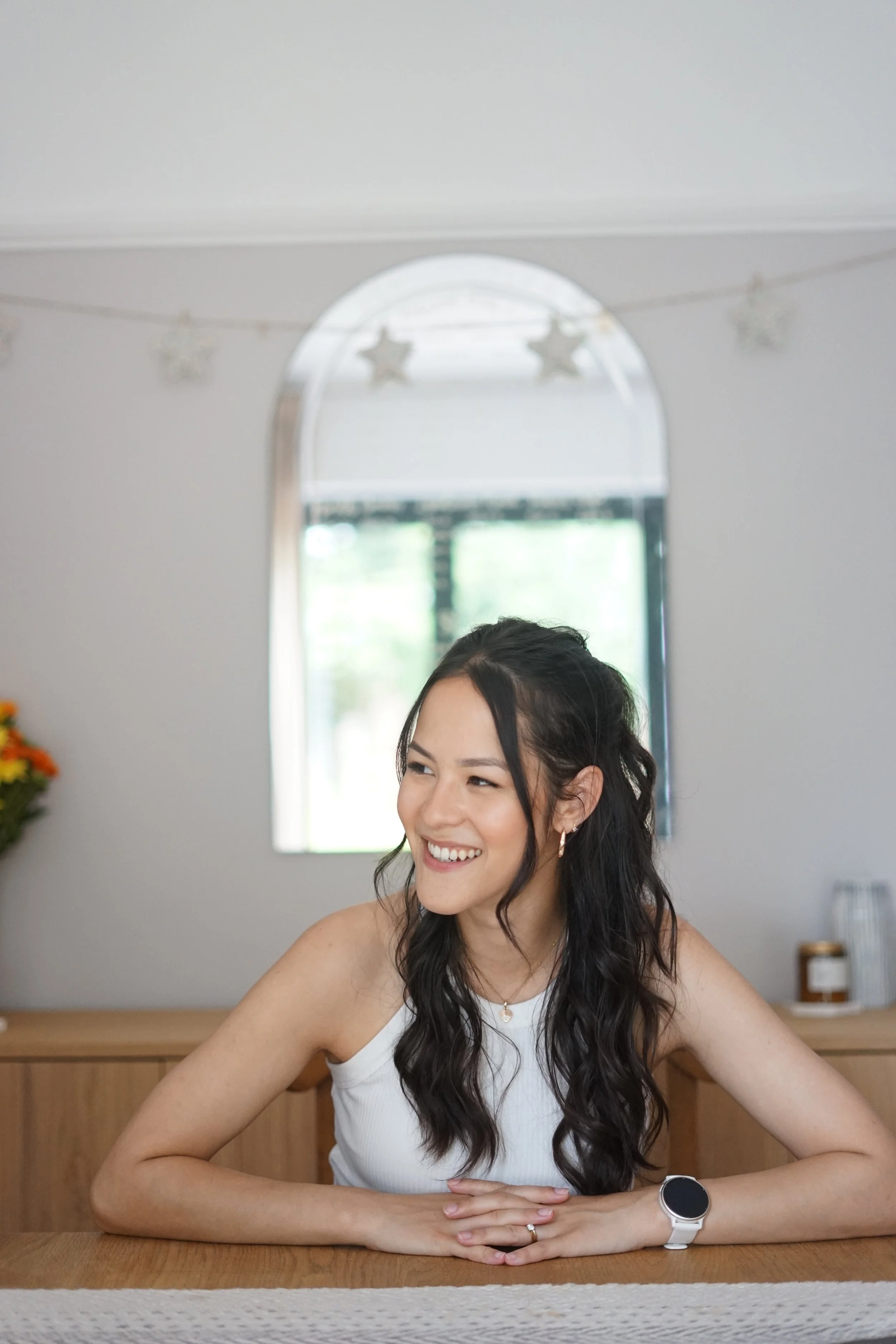 A woman sitting at a wooden table smiling, with dark wavy hair, wearing a white tank top, a wristwatch, a necklace, and a ring, in a room with a mirror, star decorations, and a window with greenery outside.