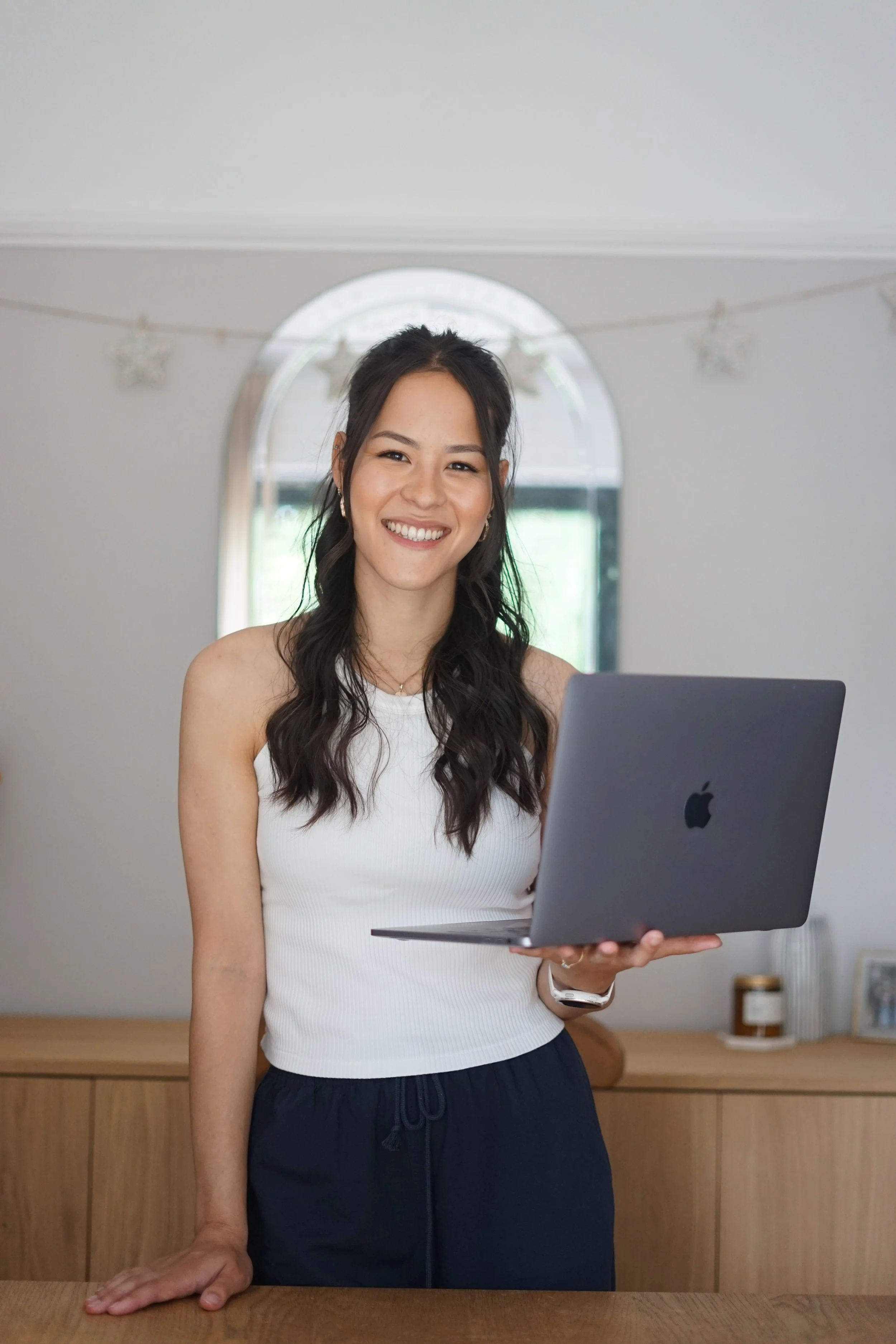 A woman with long dark hair smiling, holding a silver laptop in her right hand, standing in a room with a wooden table and a cabinet in the background.