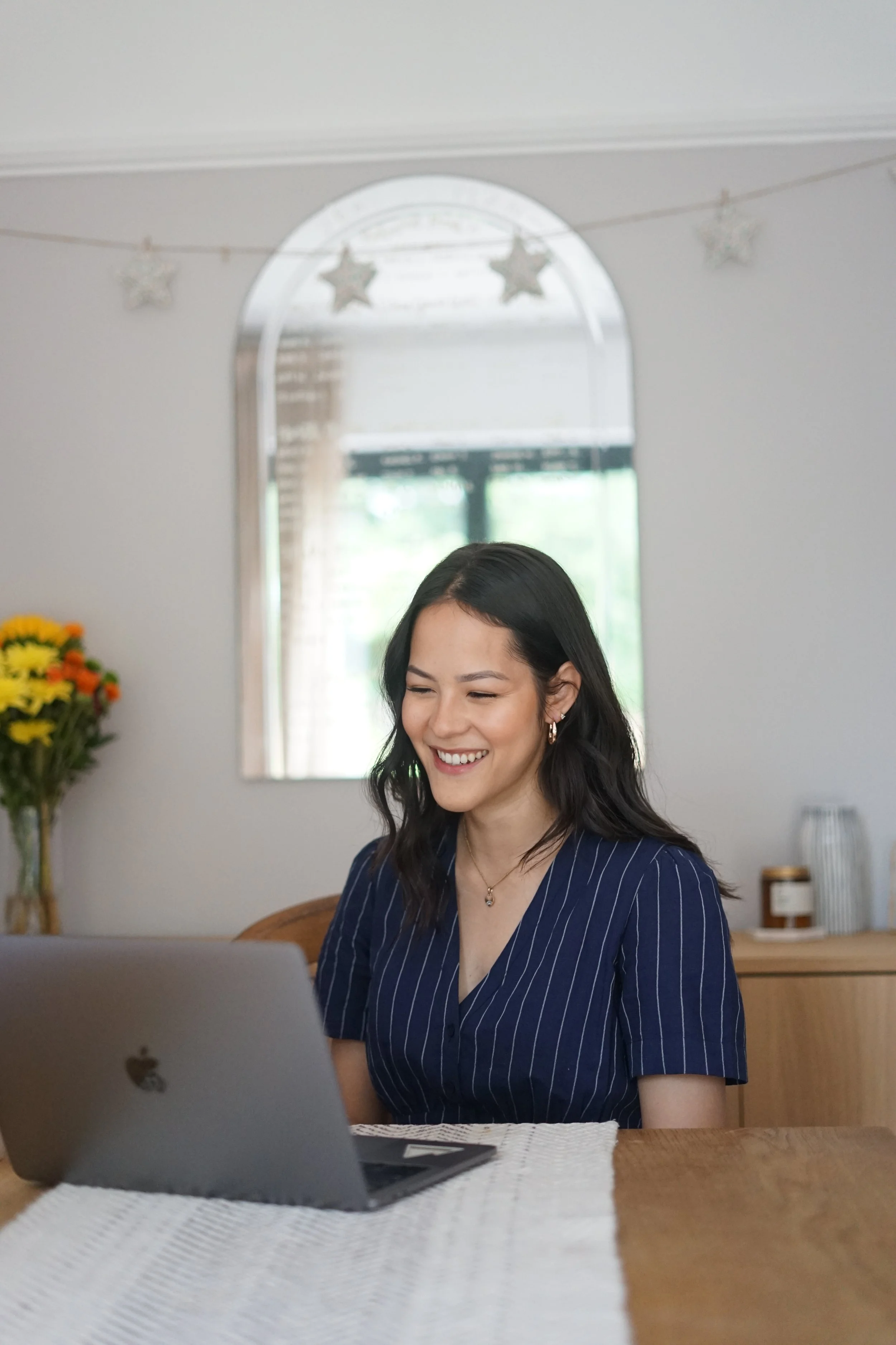 A woman with long dark hair, wearing a navy blue pinstriped shirt, smiling and working on a laptop at a wooden table in a bright room with natural light, a mirror with star-shaped decorations, a bunch of yellow flowers, and some jars on a sideboard in the background.