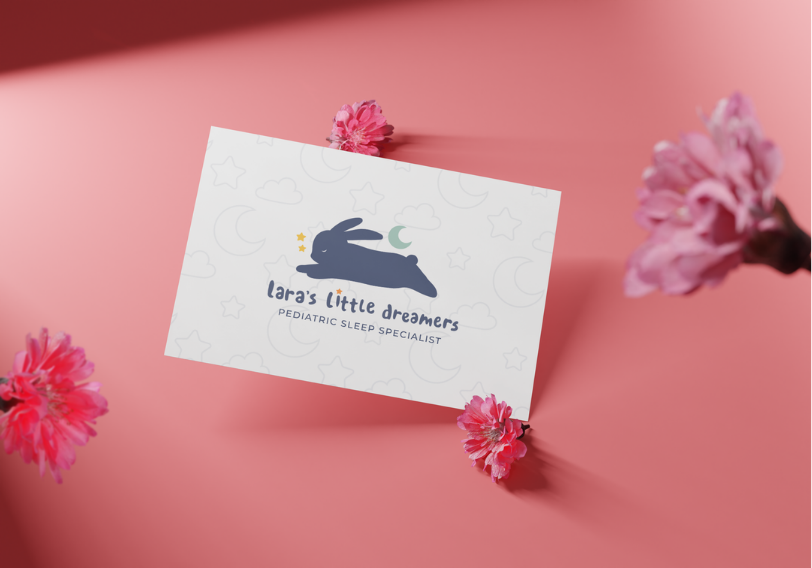 Business card for Lara's Little Dreamers Pediatric Sleep Specialist, placed on a pink surface with pink flowers around it.